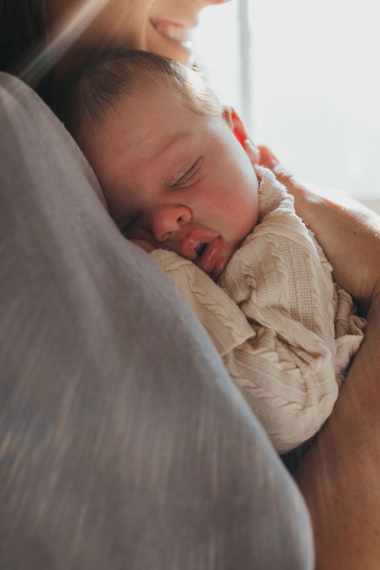 Sleeping infant nestled against soft gray fabric in window light. Wichita newborn photographer in-home session