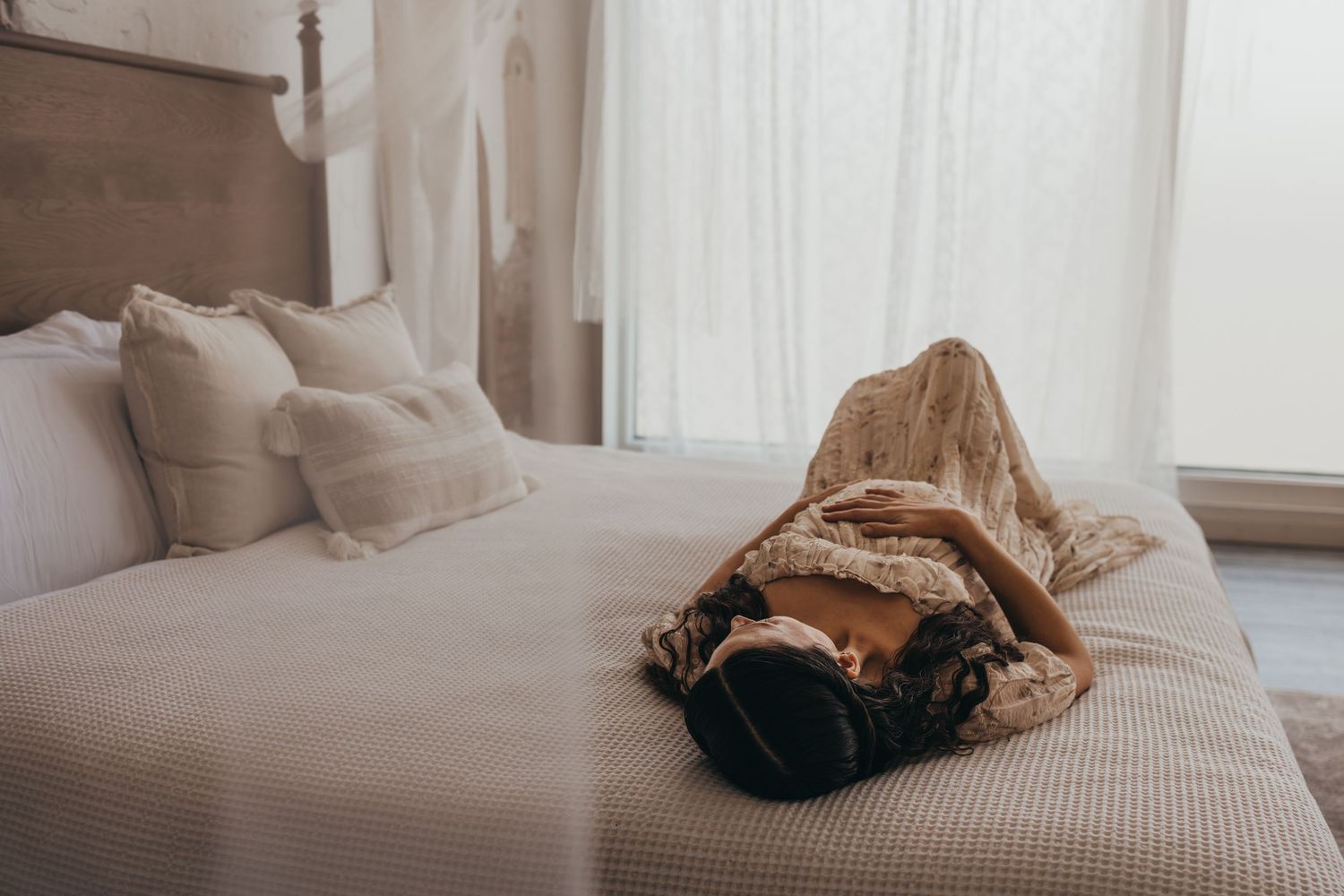 Person lying on a cozy bed with white linens and a brown blanket next to sheer curtains.