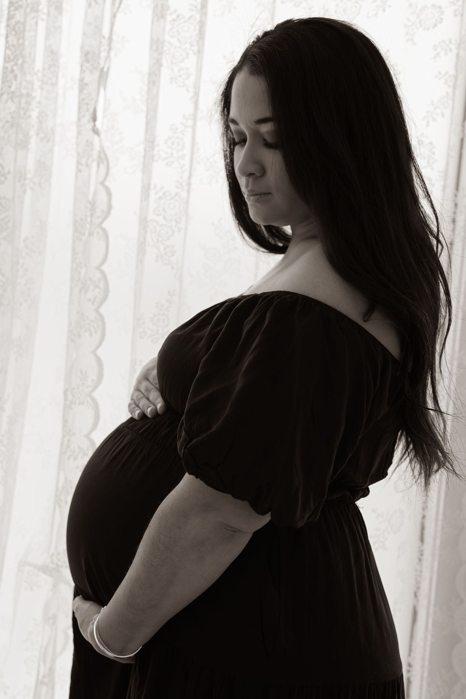 Black and white maternity portrait in off-shoulder dress by window.
