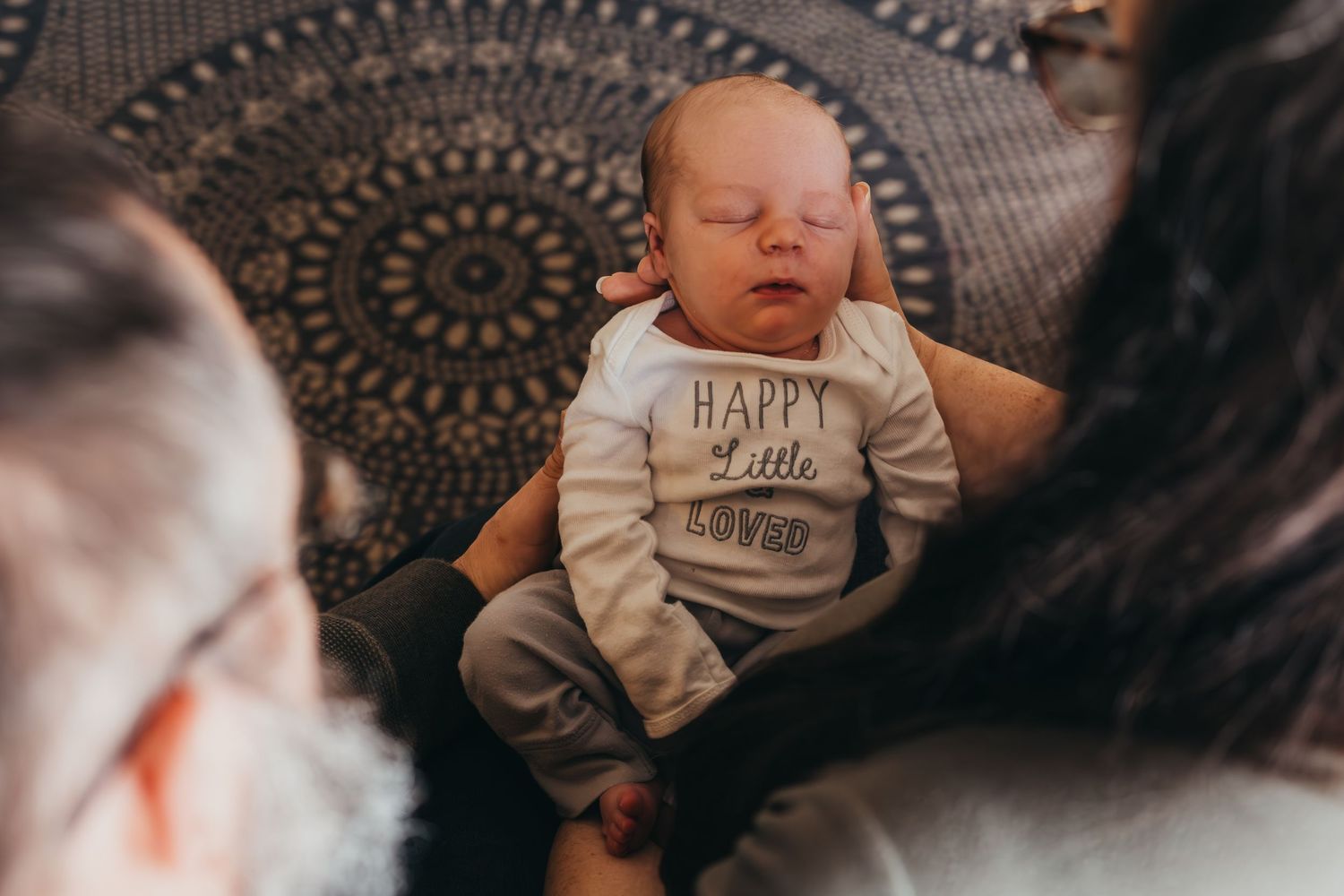 A baby wearing a white onesie lies on a patterned blanket while being photographed from above.
