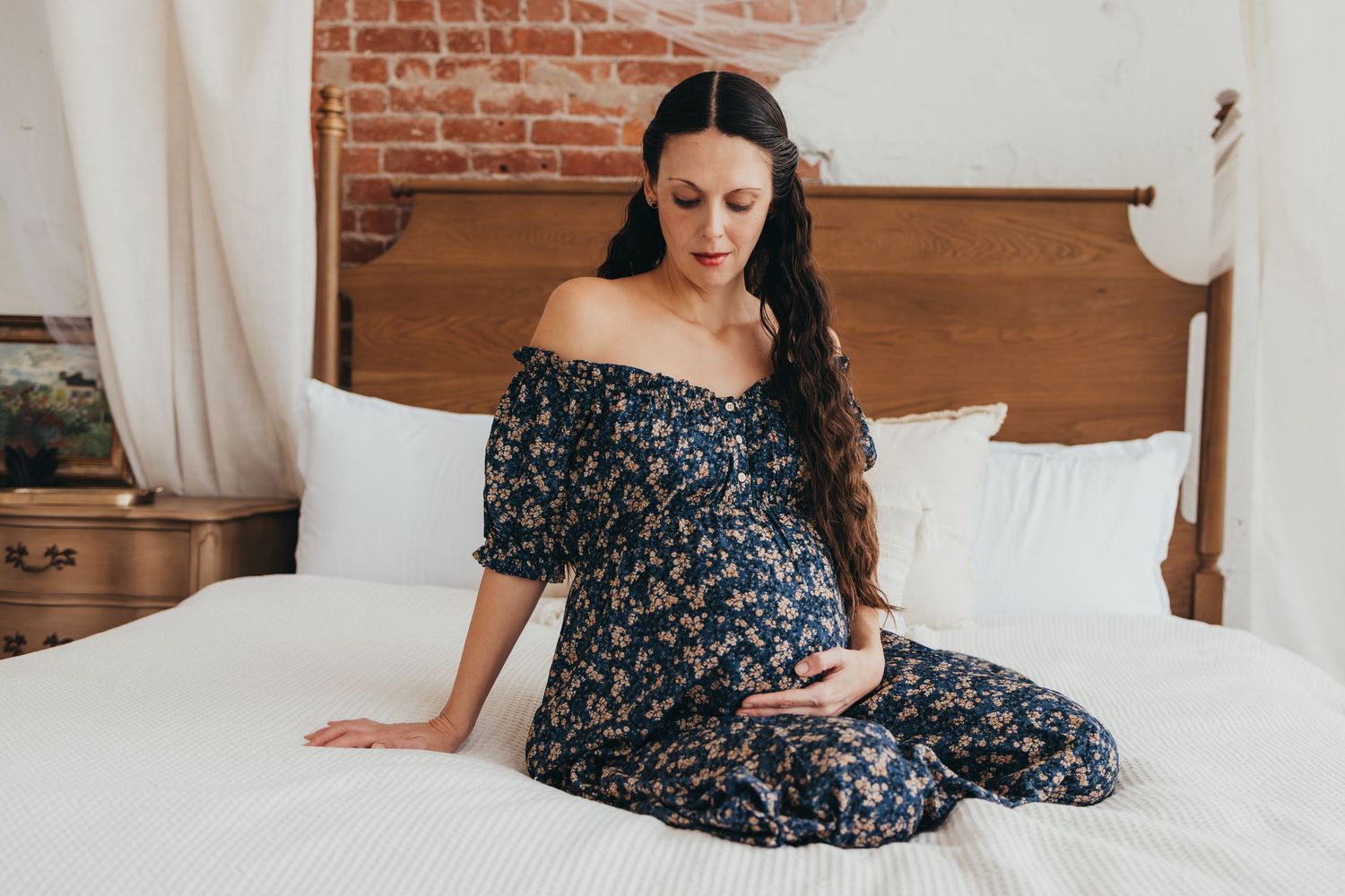 Expectant mother in floral dress sitting on white bed with wooden headboard.