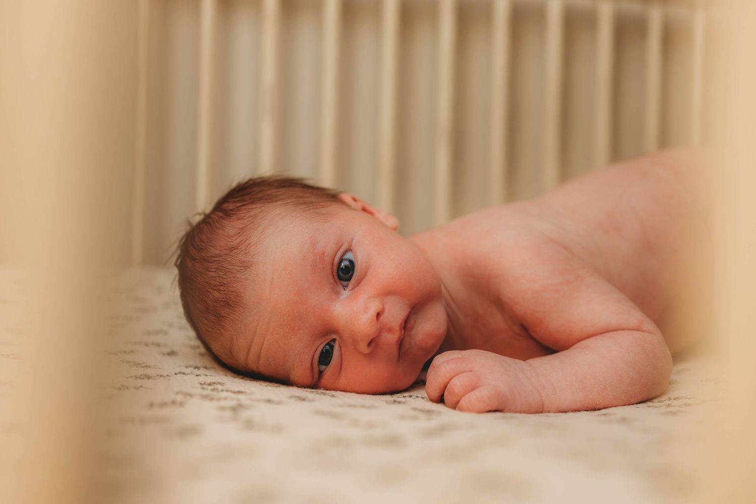 Infant lying peacefully on cream colored bedding in natural window light.