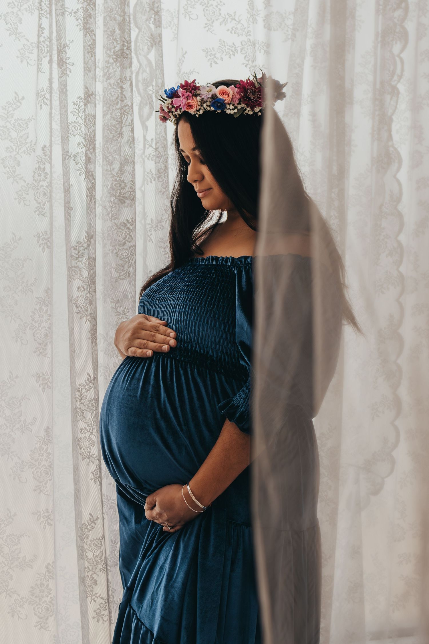 Maternity portrait with floral crown and blue dress against white curtains.