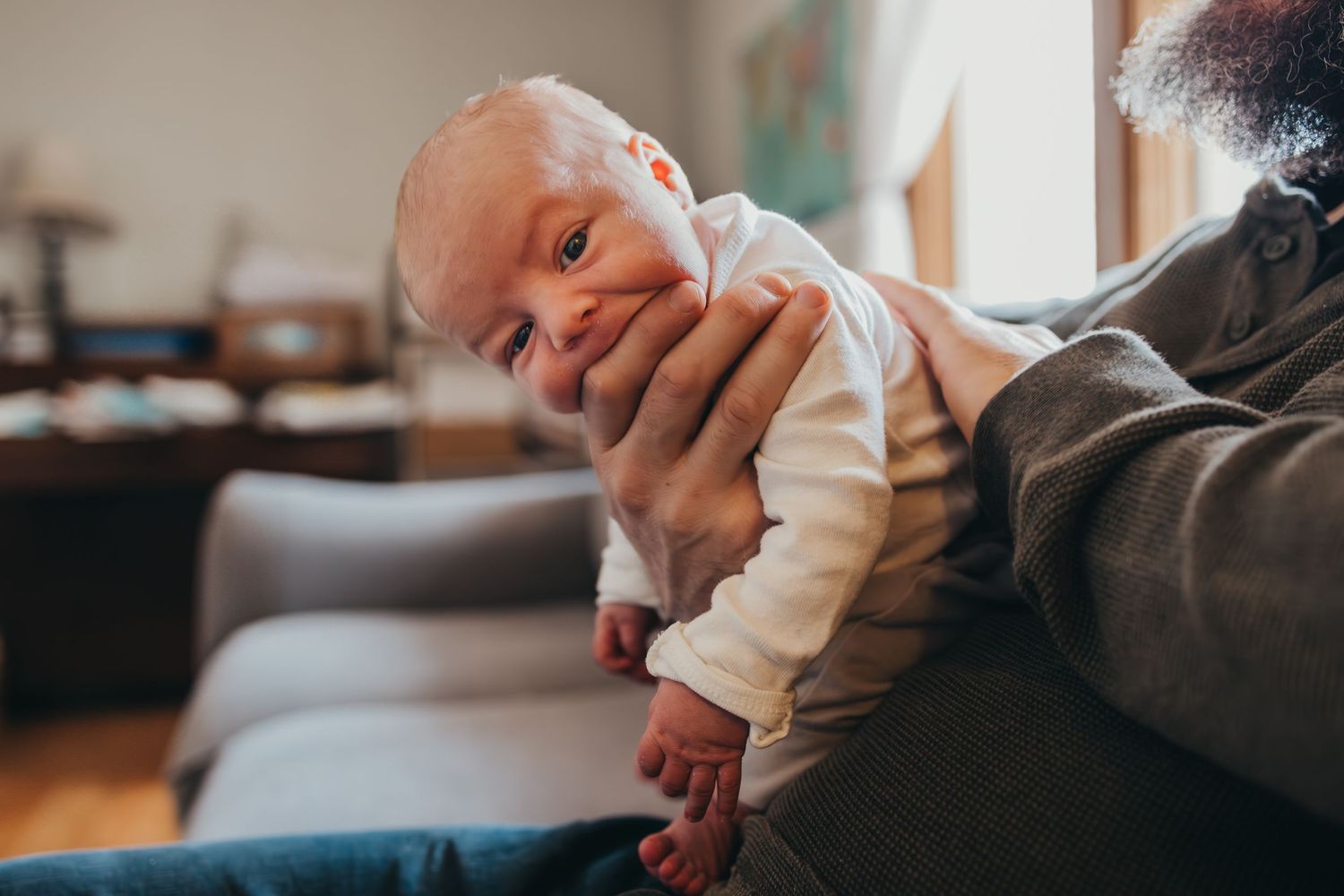 A baby in a cream-colored onesie lies on a couch while being comforted by someone's hands.