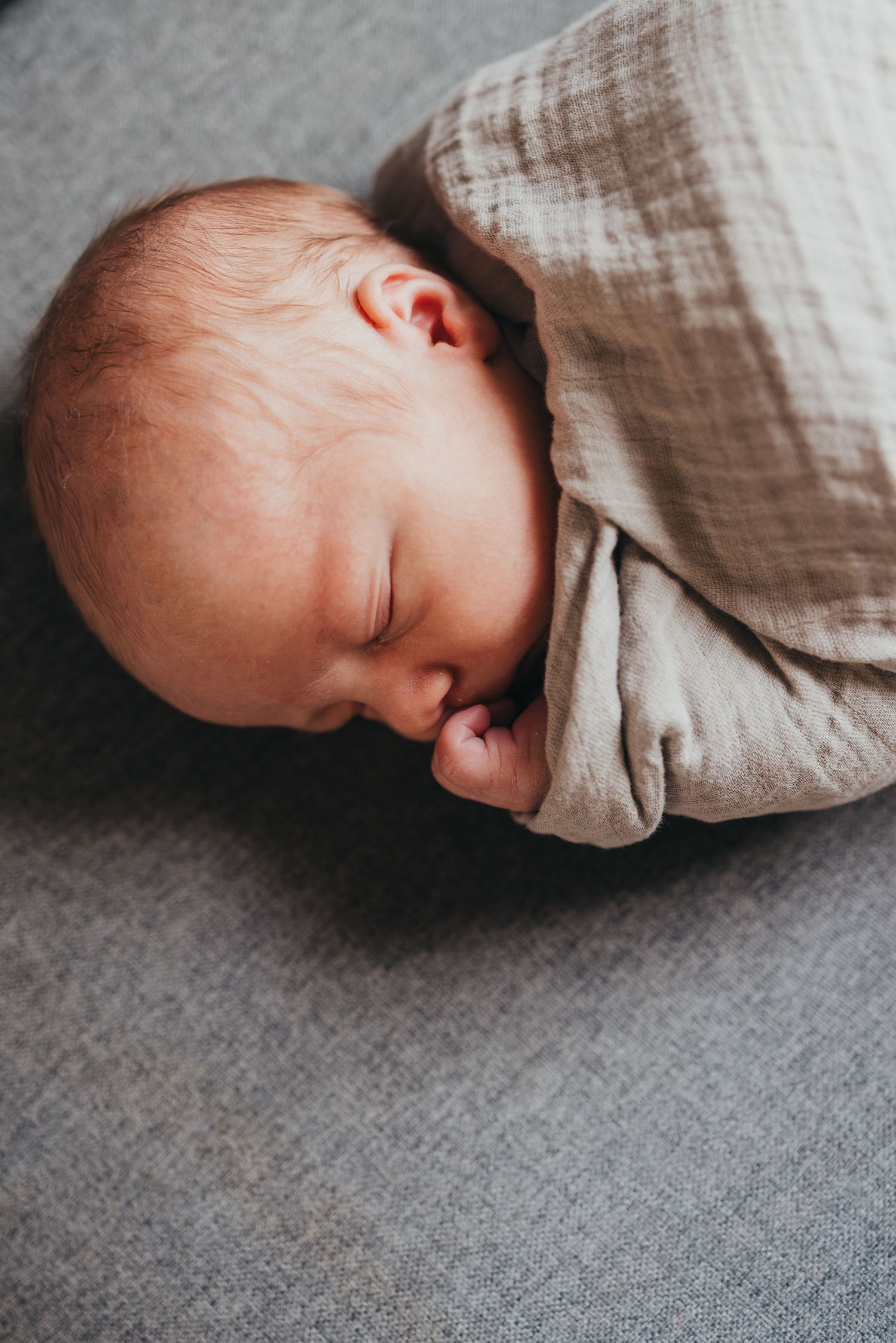 A sleeping baby lies peacefully on a grey textured surface wrapped in soft beige linen fabric.