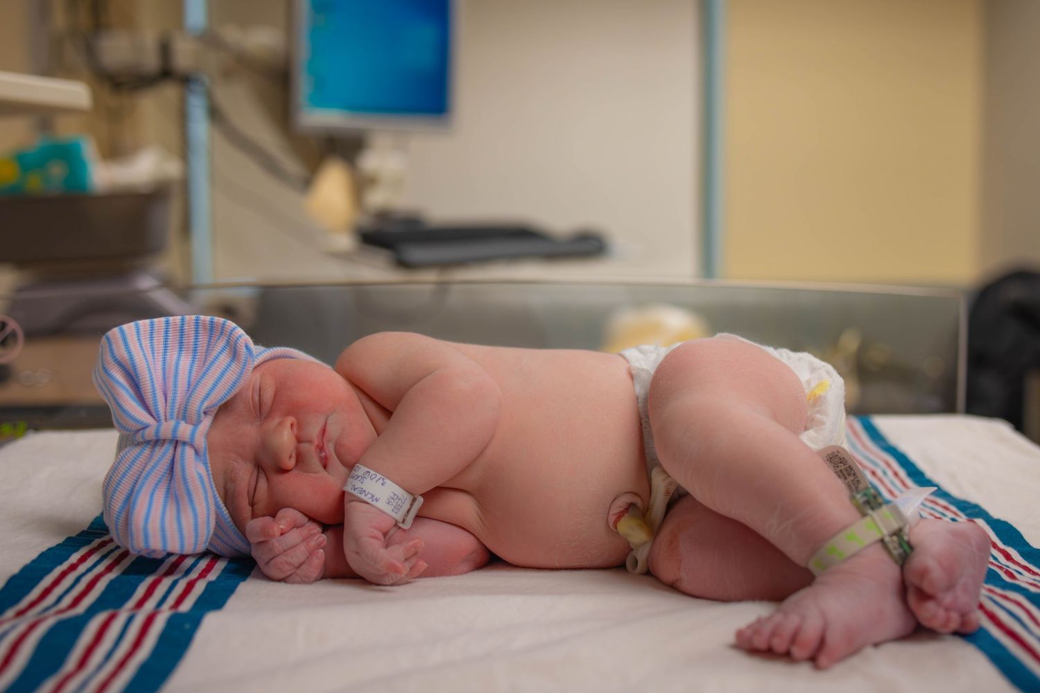 A sleeping newborn baby wearing a striped hospital hat lies on white hospital bedding.