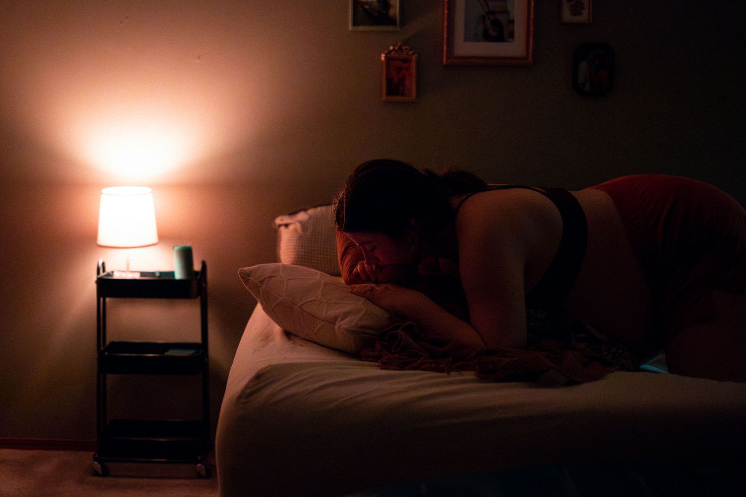 A person lies on a bed in a dimly lit room with a warm bedside lamp glowing in the background.