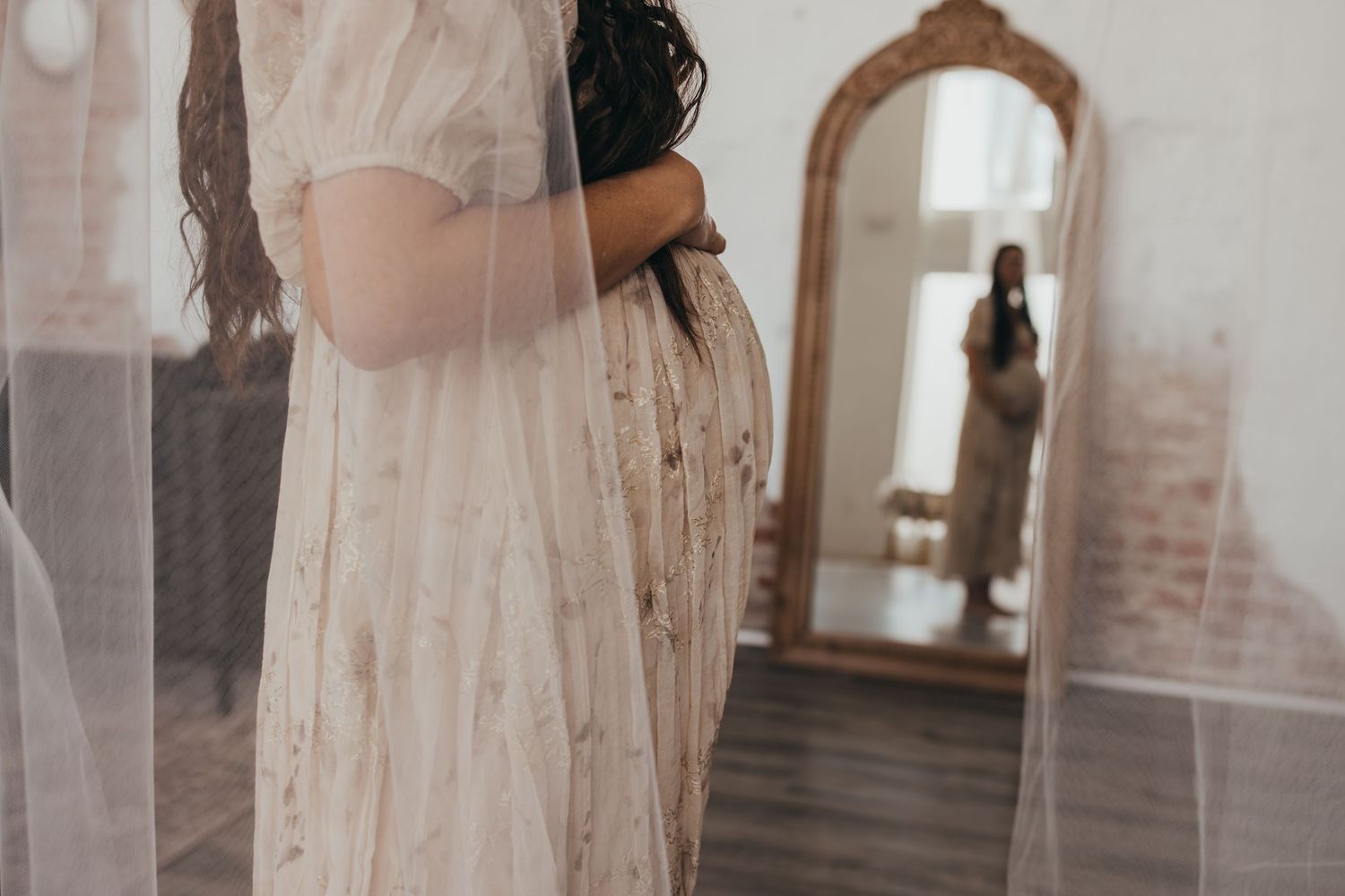 Ethereal maternity photo with flowing white dress and vintage mirror in rustic room.