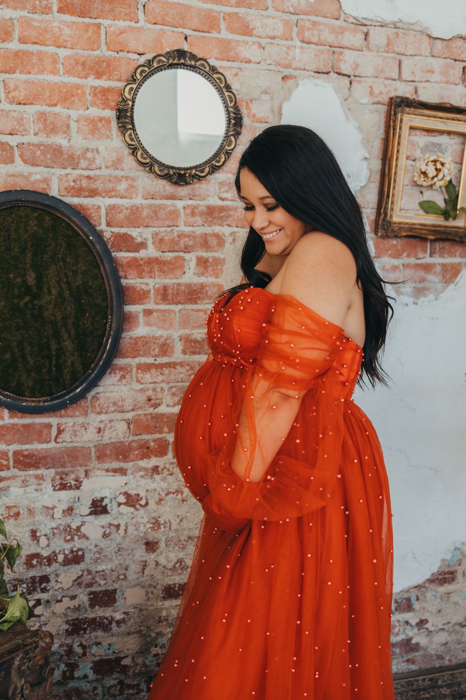 Woman in flowing orange dress poses against rustic brick wall with vintage mirrors.
