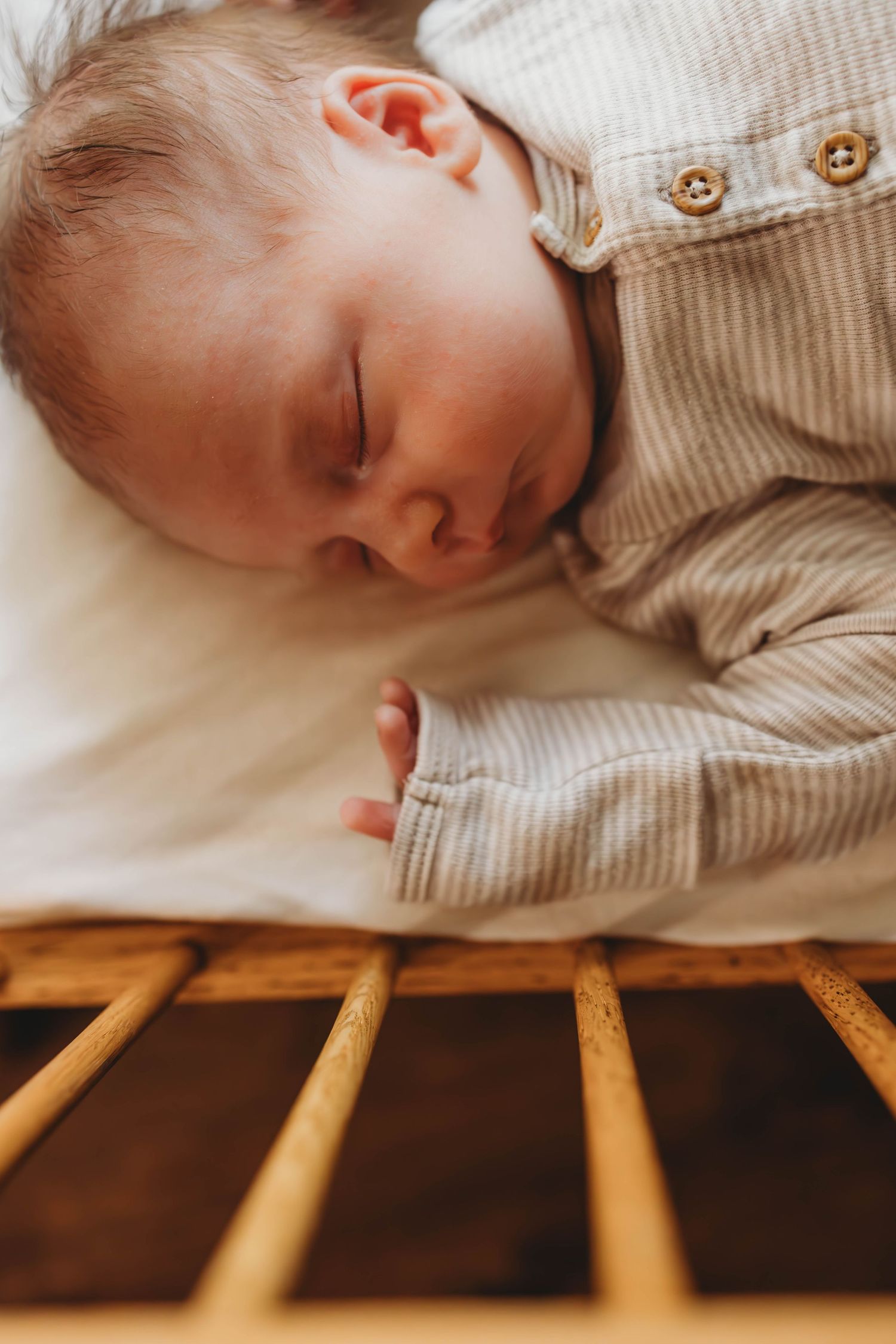 Baby in striped onesie sleeping soundly on white bedding near wooden crib rails.