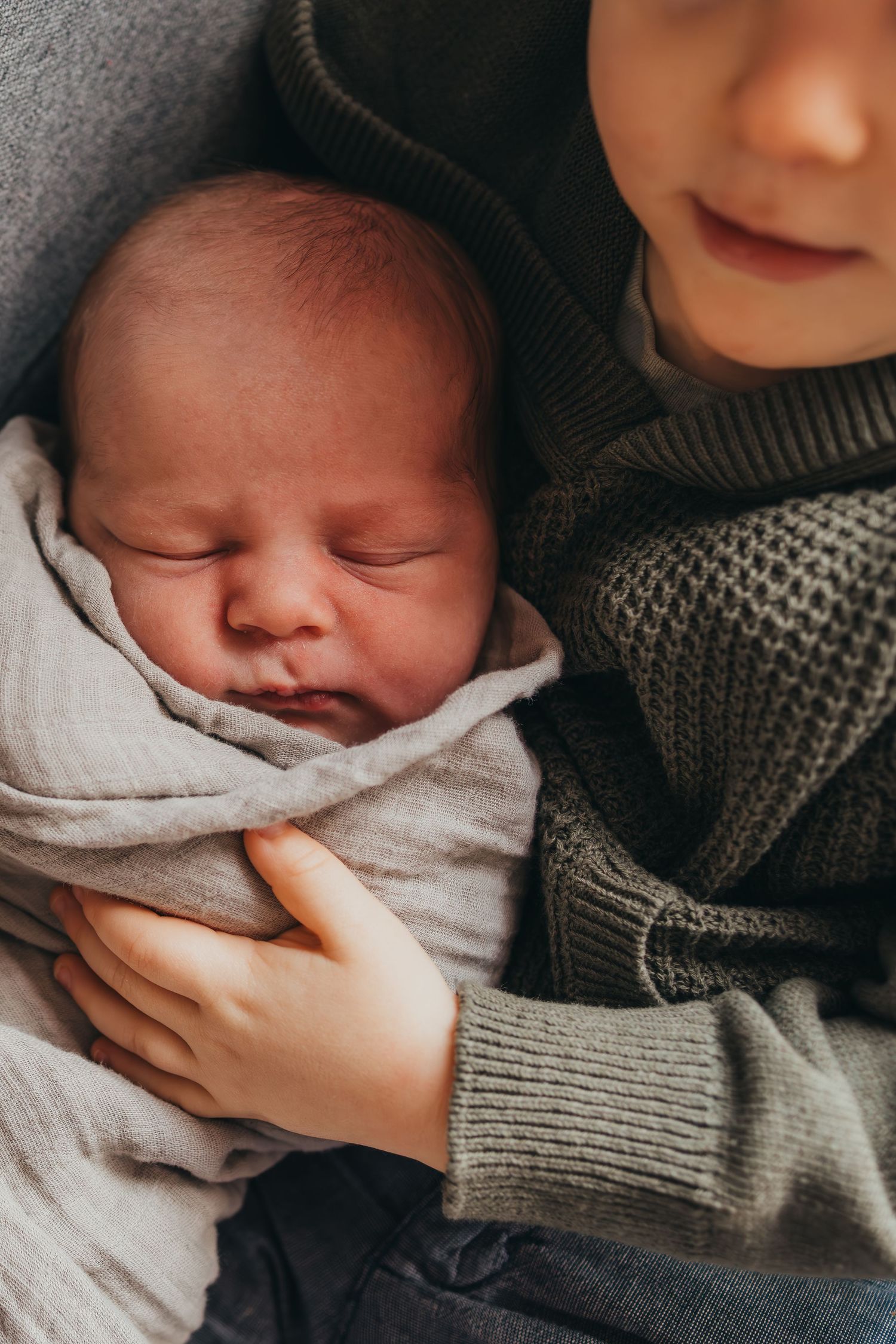 A sleeping newborn baby swaddled in light fabric is held gently in someone wearing a gray knit sweater.