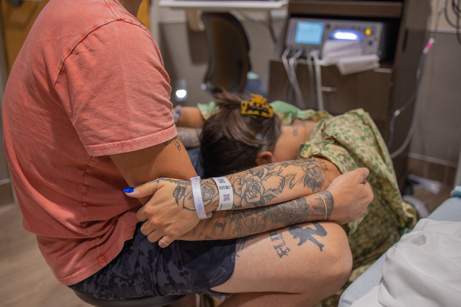 A person in a coral shirt with tattoos holds a baby in a hospital setting.