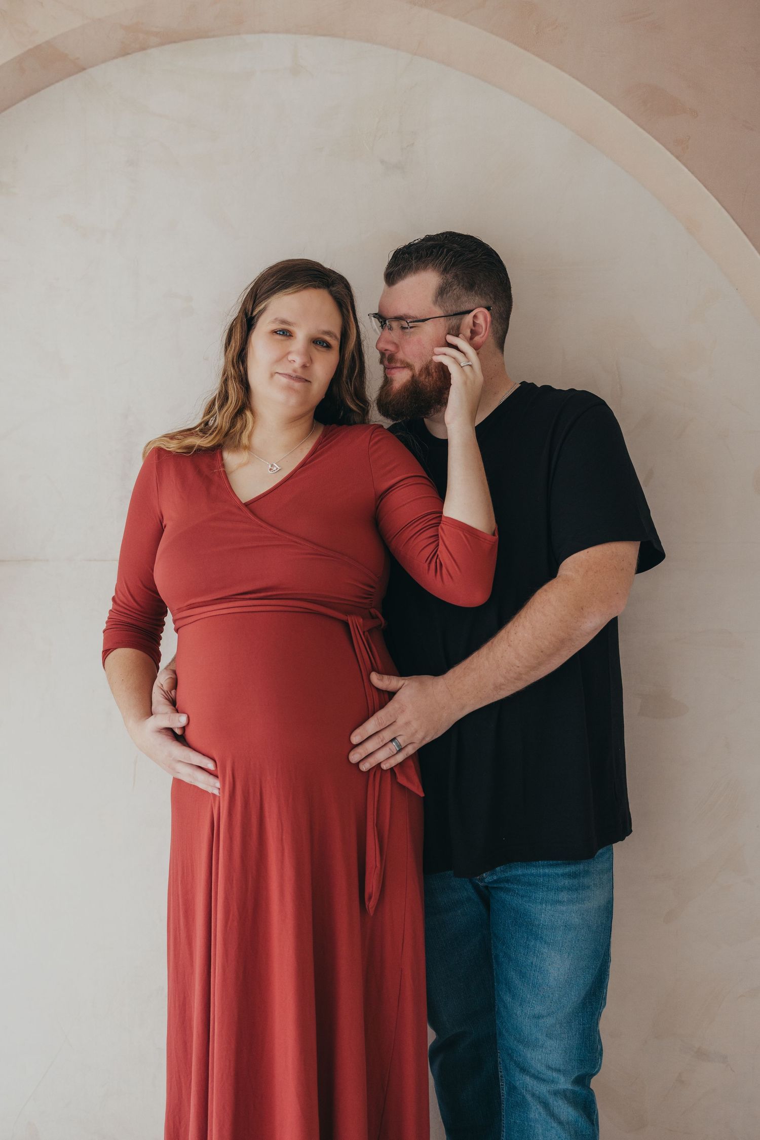 Couple in casual attire poses against neutral wall, person wearing red wrap dress and partner in black shirt and jeans.