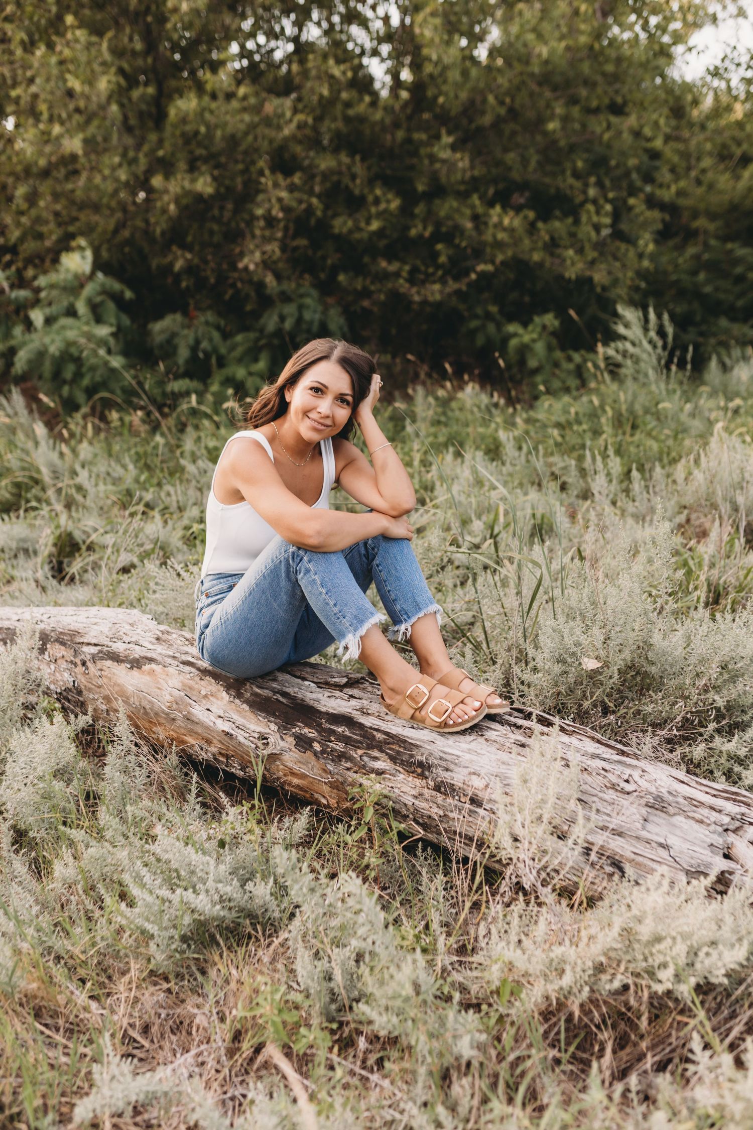 Woman sitting on a weathered log surrounded by tall grass during a sunset photoshoot.