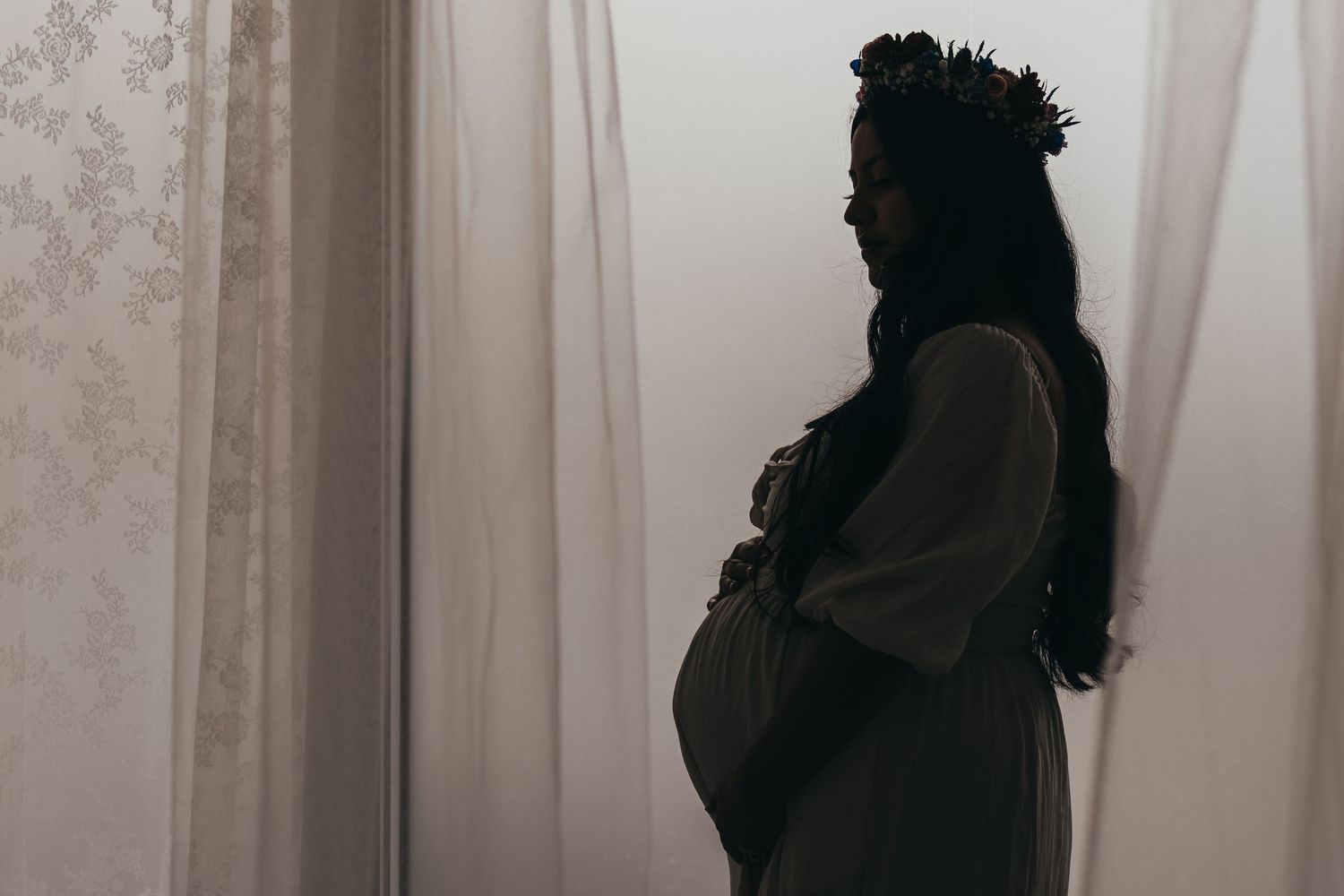 Silhouette maternity photos against white curtains in natural window light.