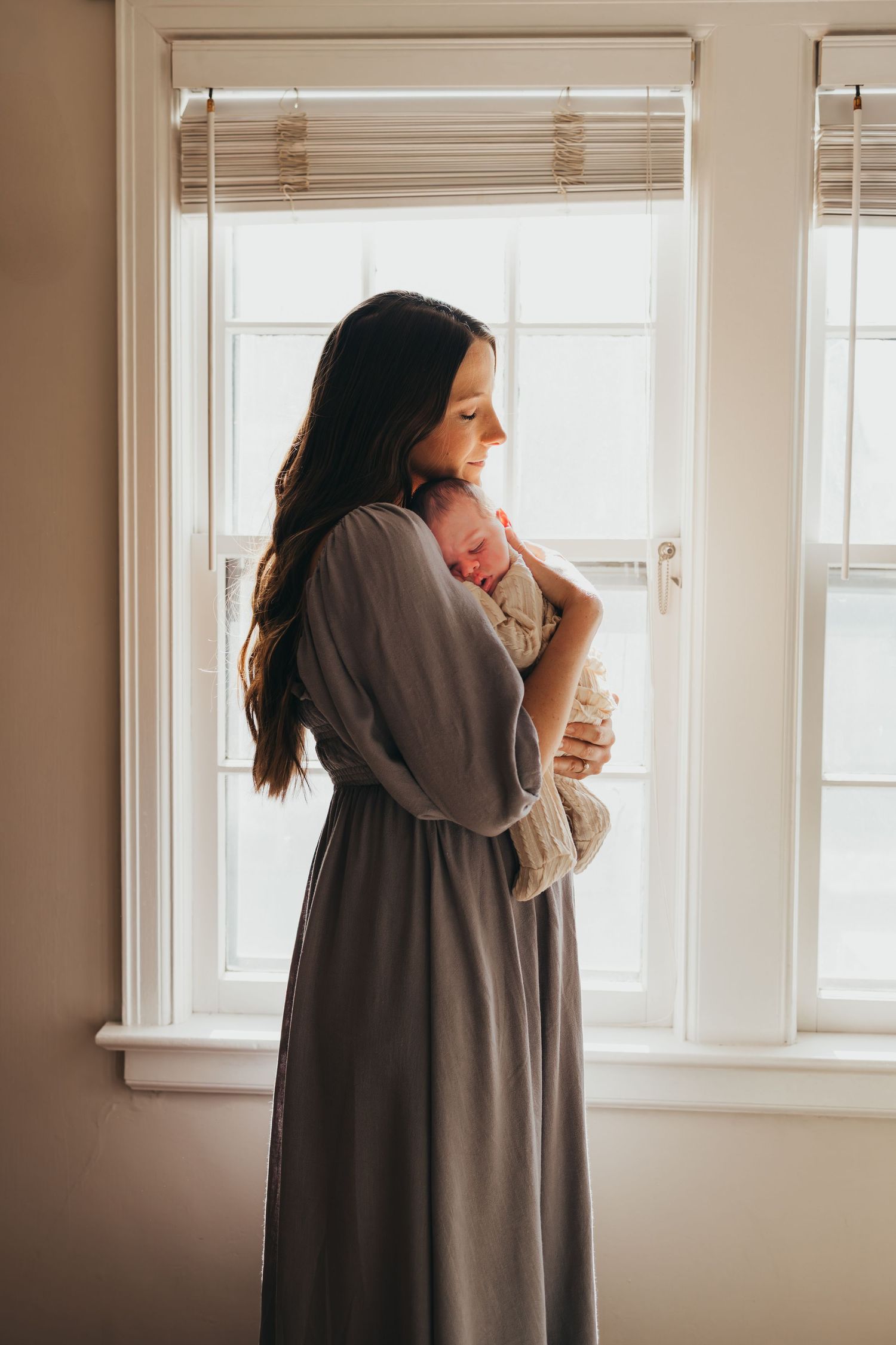Person in gray sweater holding baby near bright window. Wichita newborn photographer in-home session
