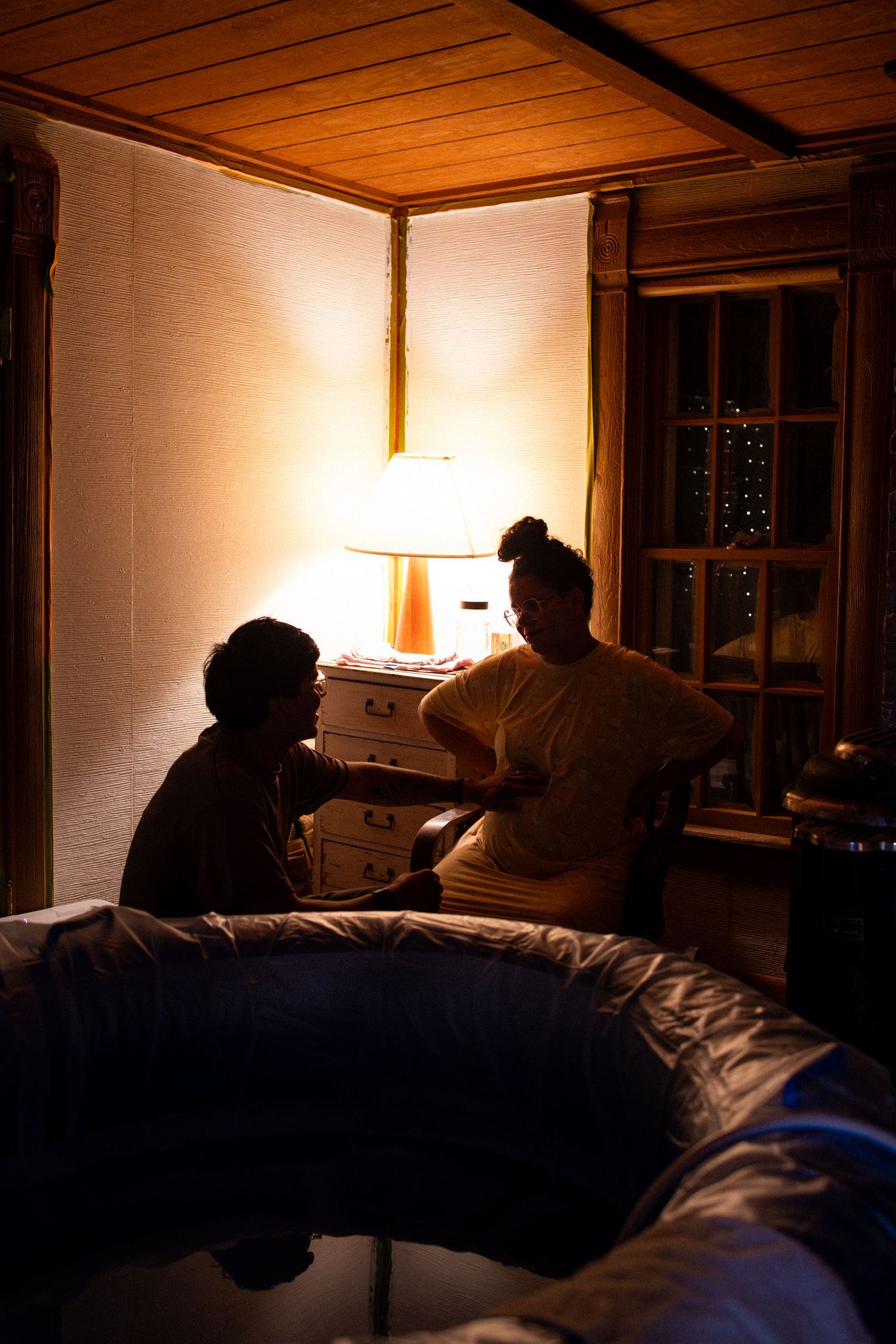 Two people sit in silhouette by a glowing bedside lamp in a warmly lit bedroom at night.