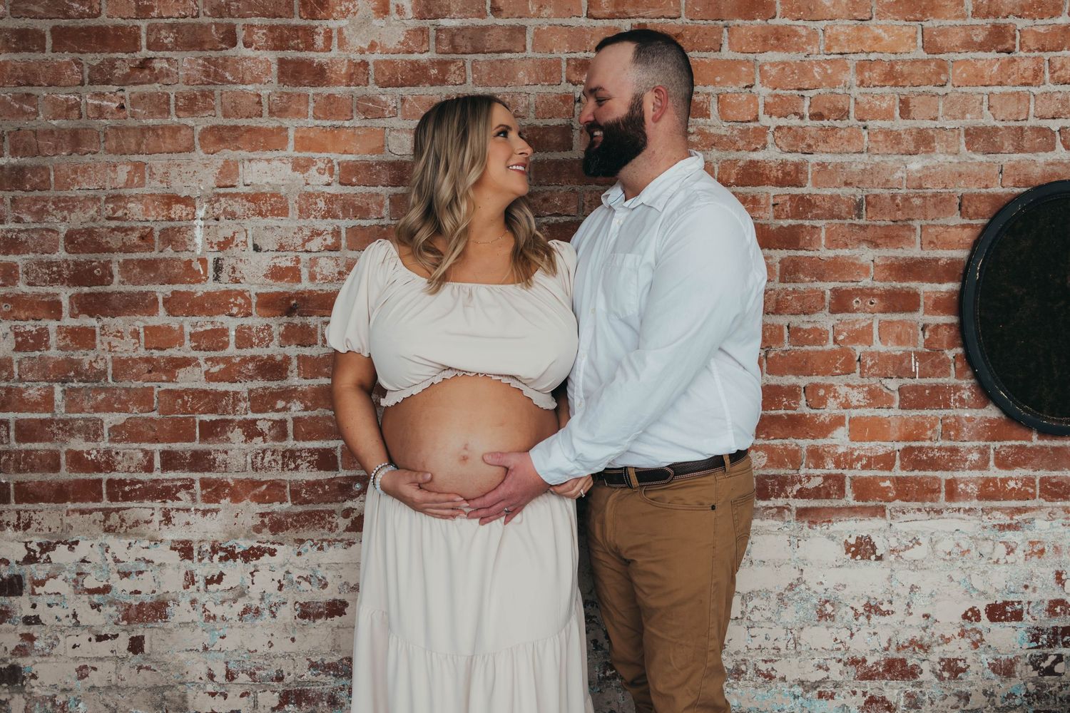 Maternity photo session against rustic brick wall showing couple in casual attire.
