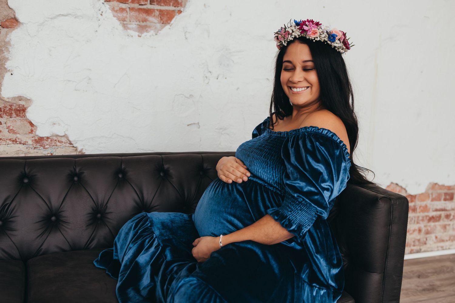 Maternity photo in blue velvet dress with floral crown on leather couch.