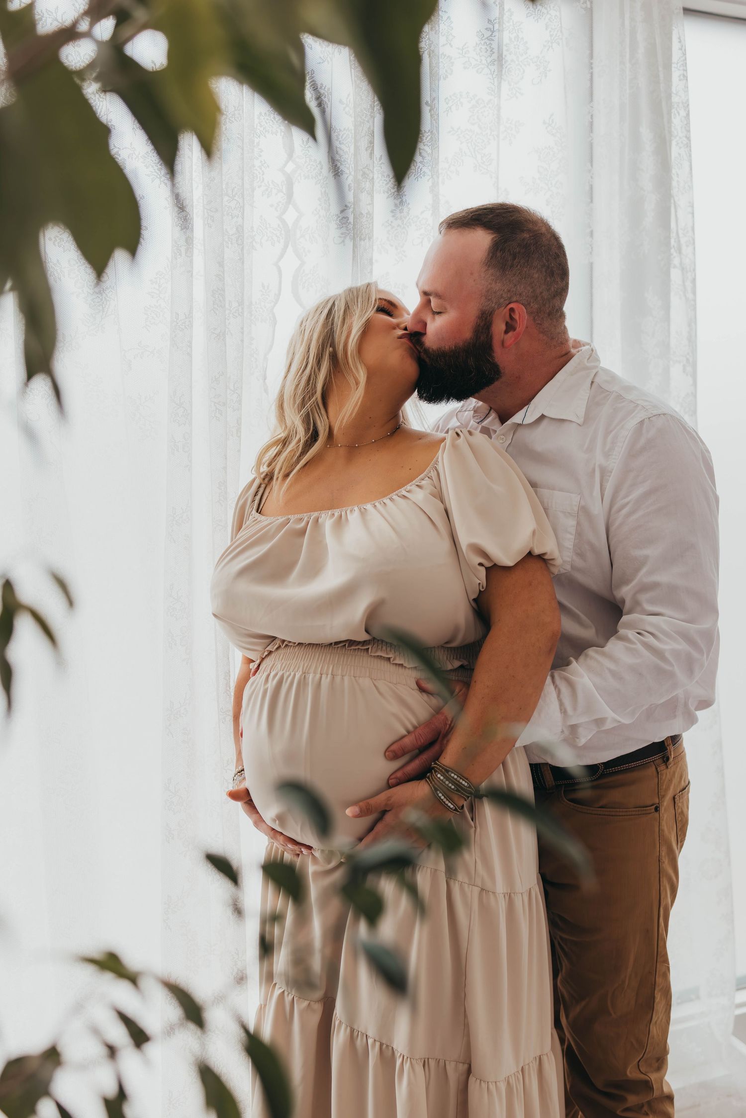 Romantic maternity photoshoot with couple embracing near white curtains and greenery.