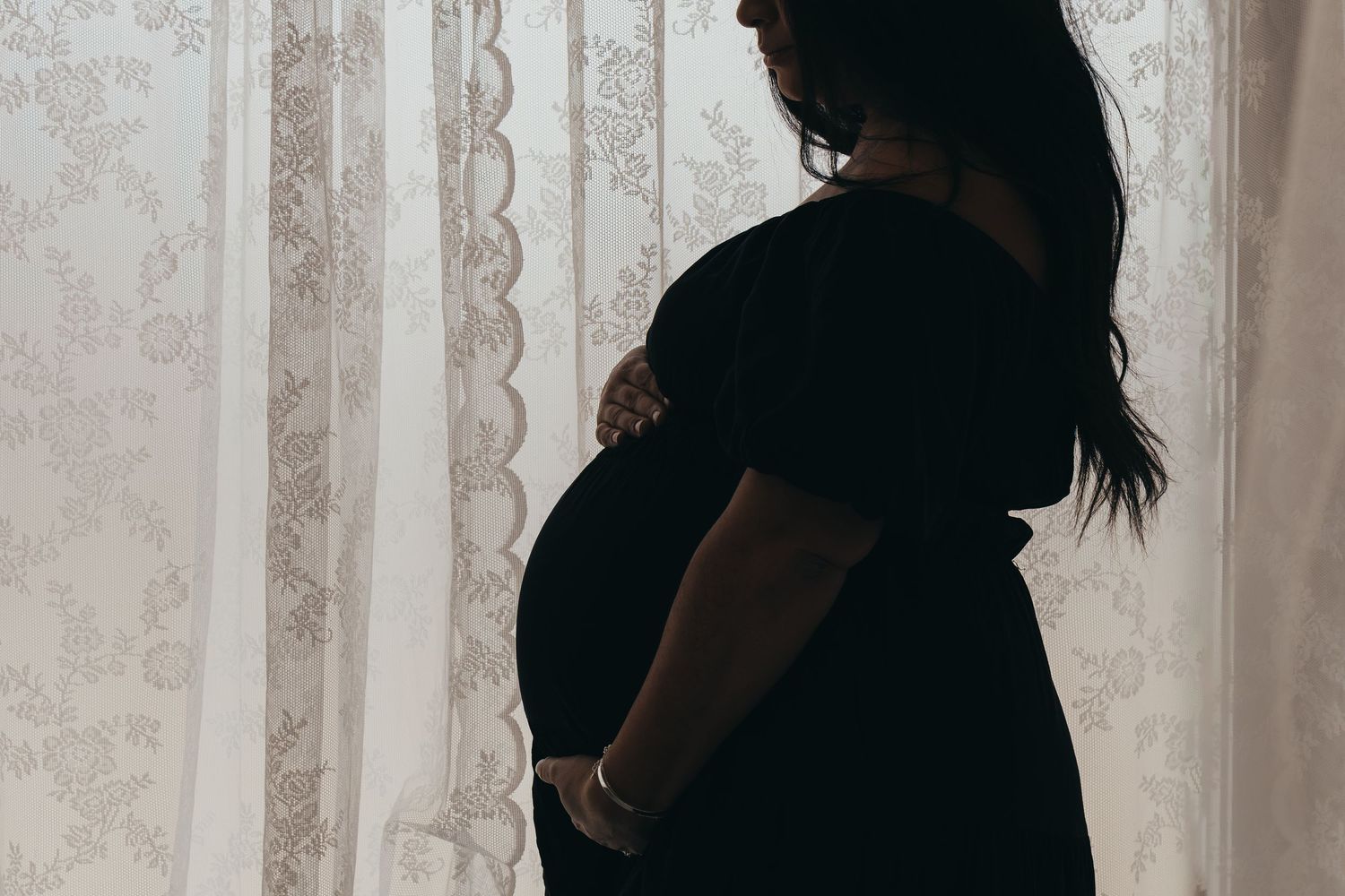 Silhouette of pregnant figure against white curtained window.