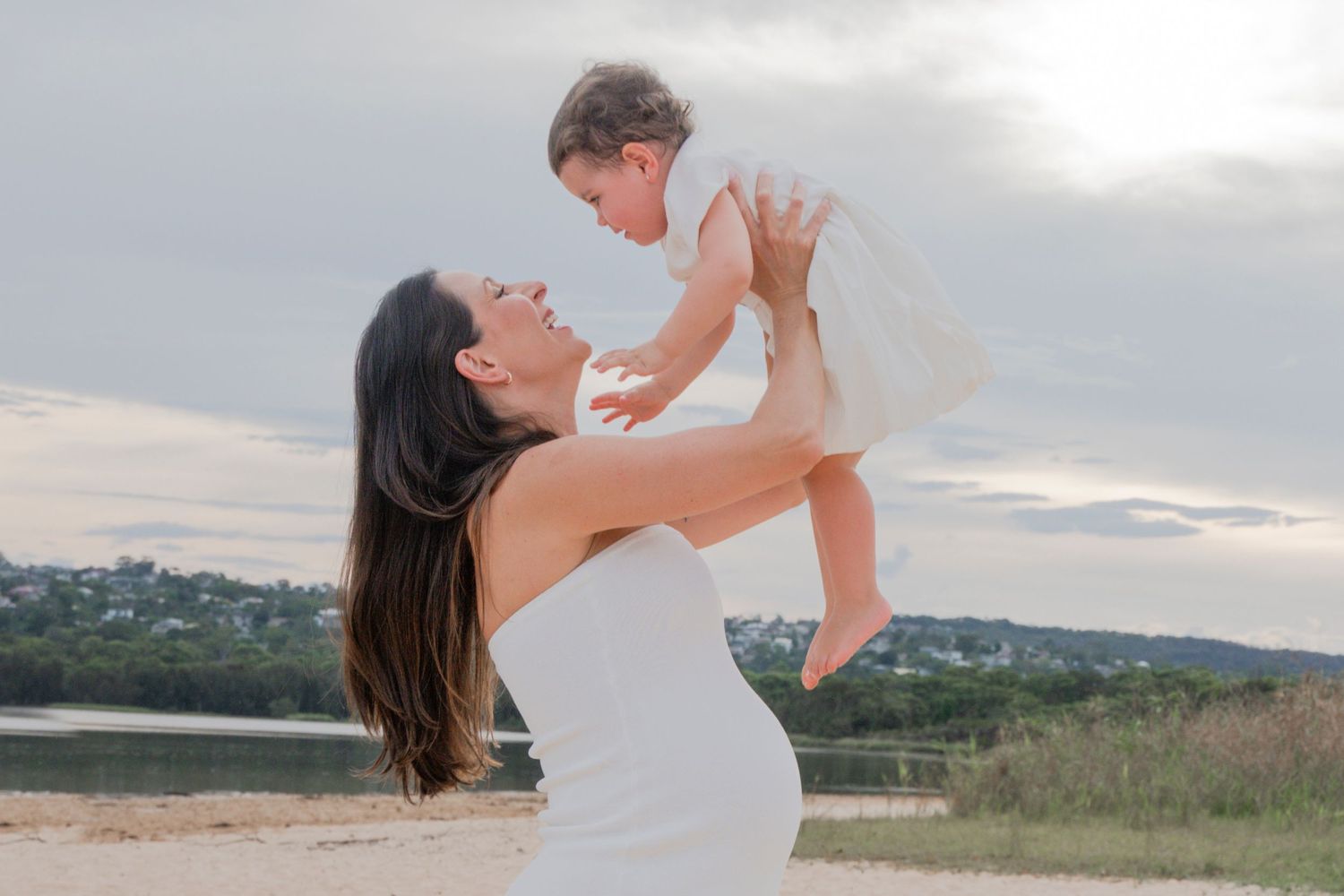 A woman in a flowing white dress lifts a baby against a scenic outdoor backdrop at sunset.