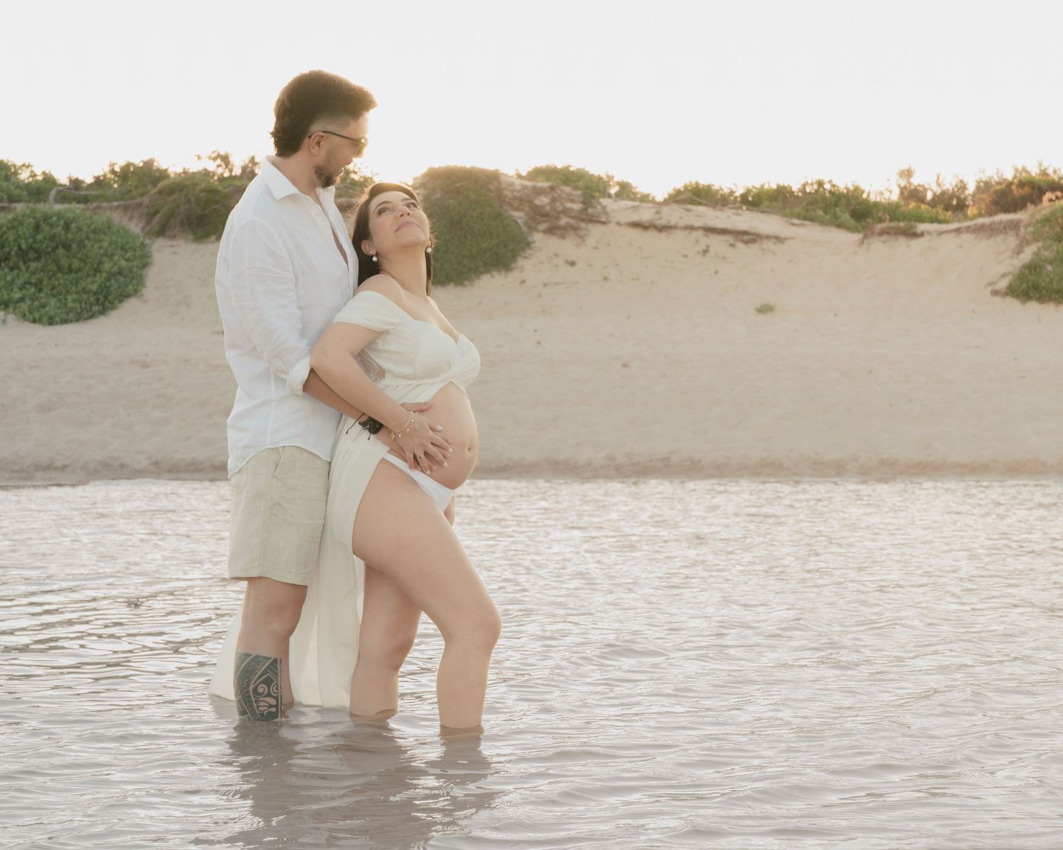Romantic maternity photo of couple standing in shallow water at sunset.