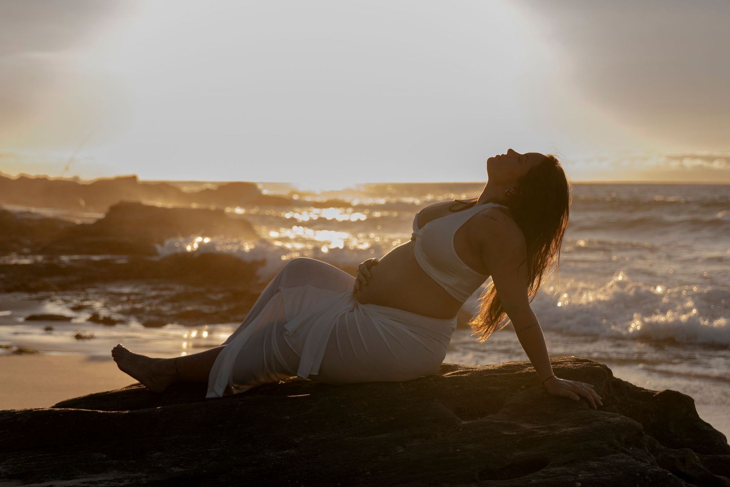 Silhouetted figures lay on beach rocks during dramatic ocean sunset.