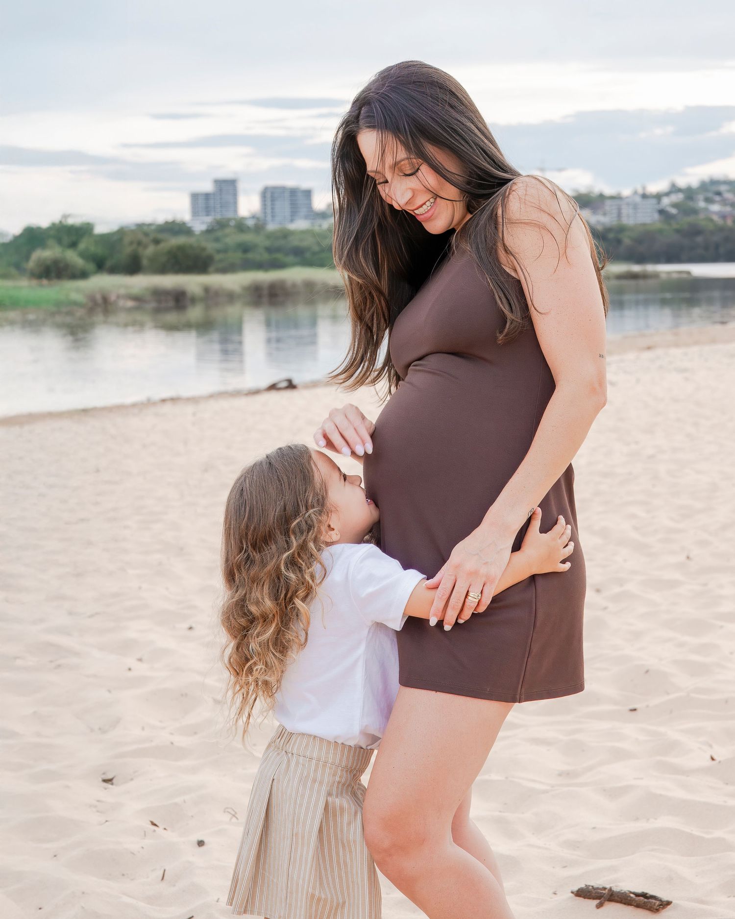 Pregnant woman in brown dress sharing moment with child on beach.