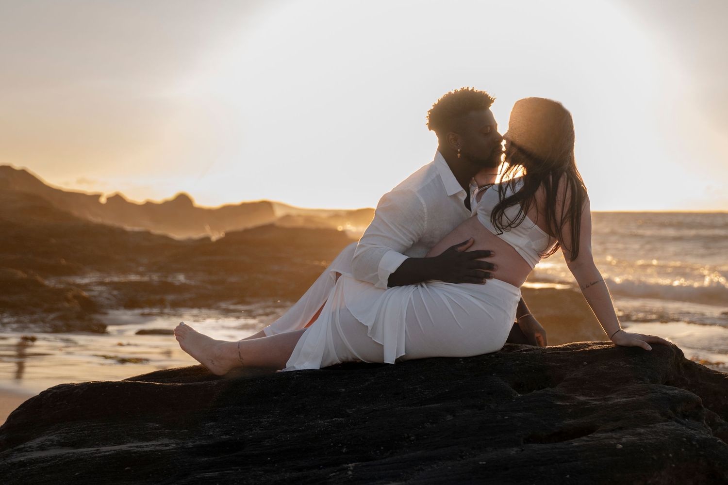 Couple shares intimate moment on rocky beach during golden hour sunset.