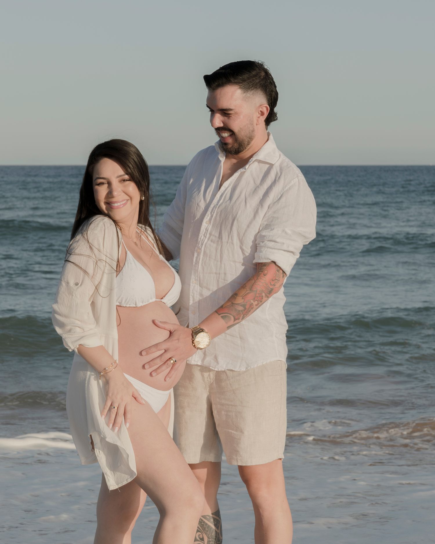 Beach maternity photo with couple in white clothing against ocean backdrop.