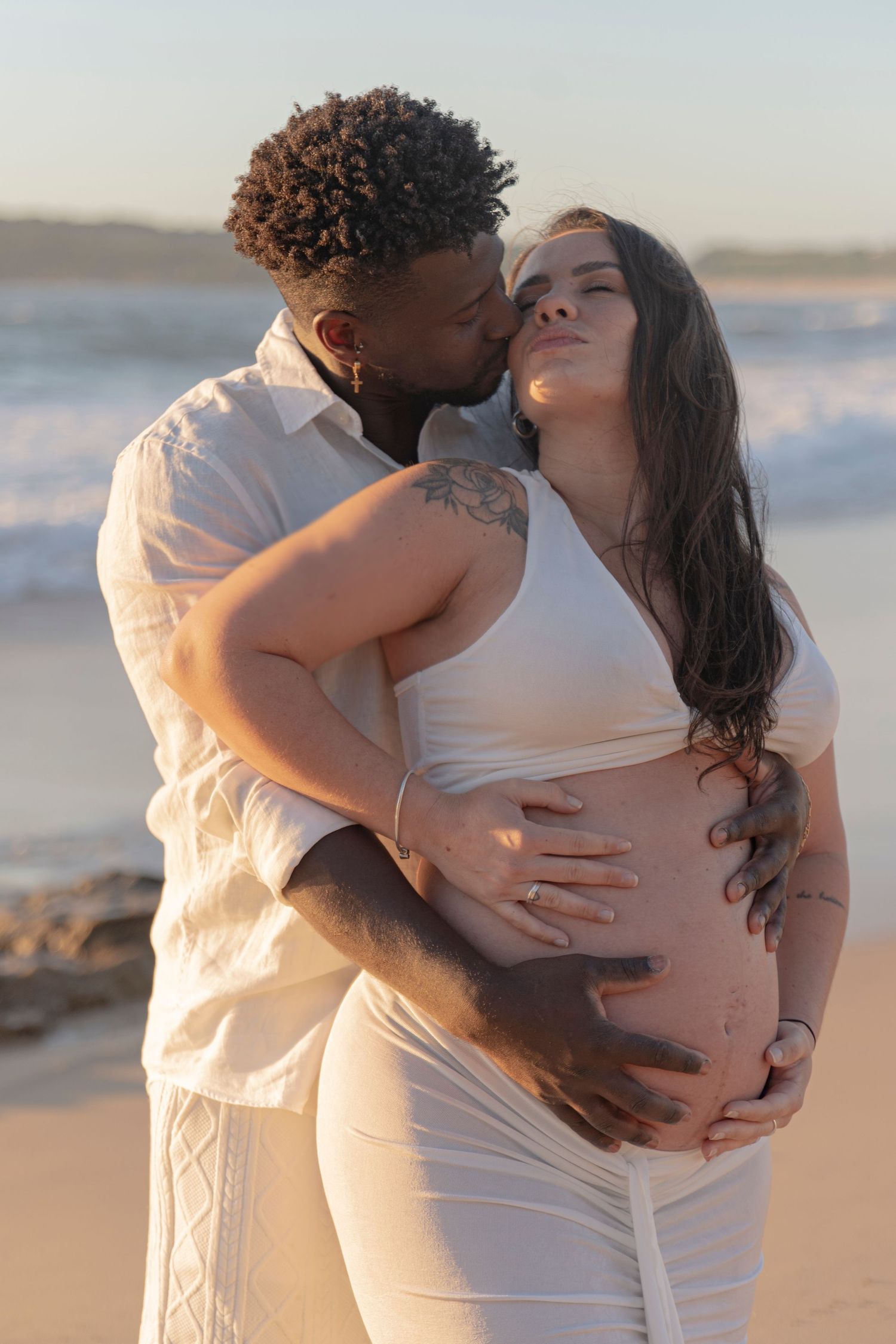 Couple in white outfits embraces tenderly on sandy beach at sunset.