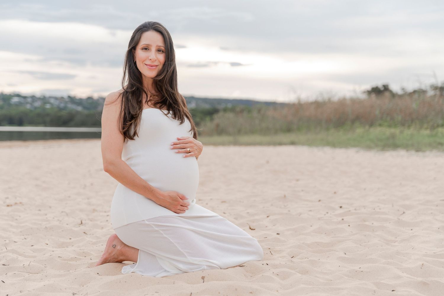 Expectant mother in white dress kneeling on sandy beach at sunset.
