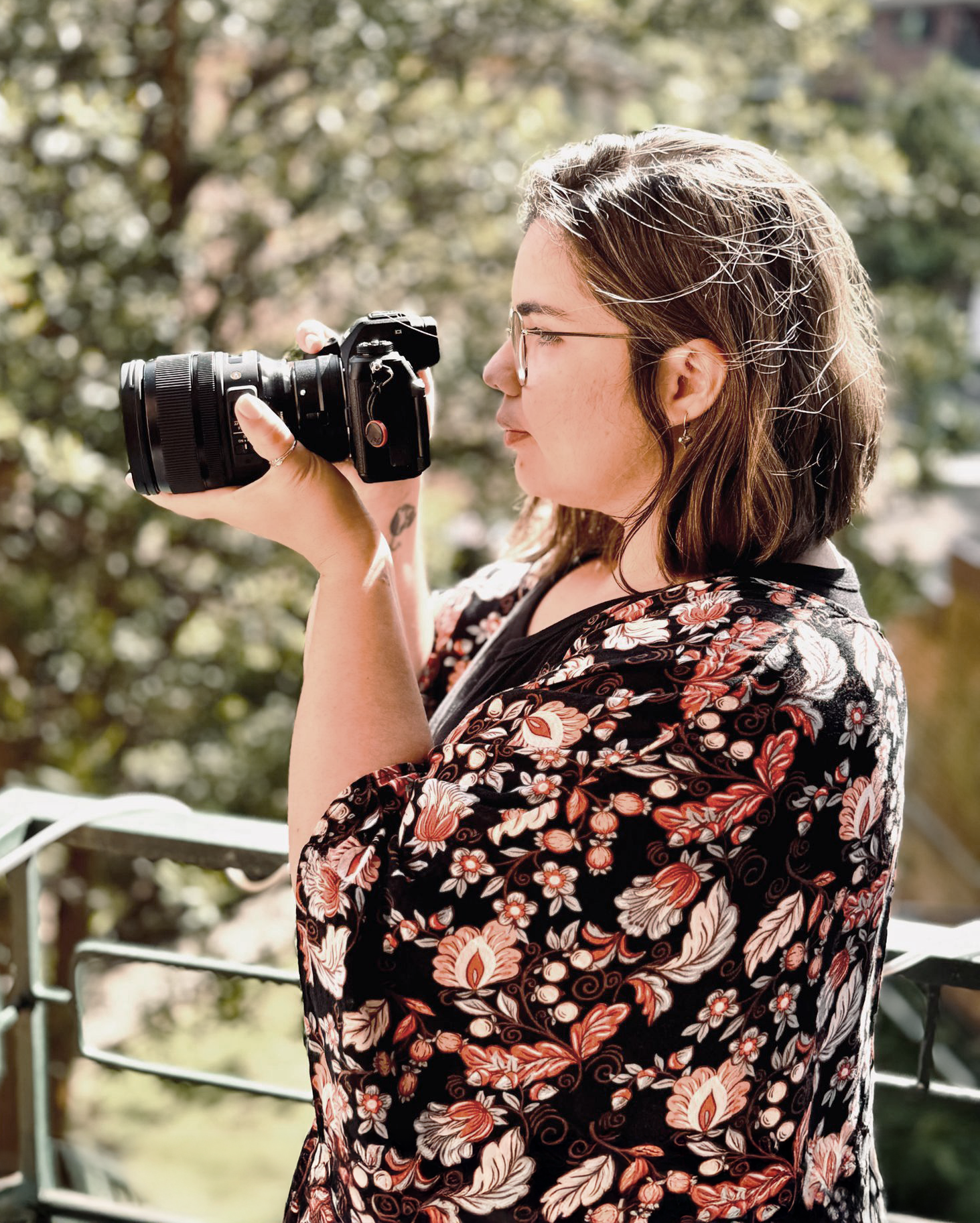 Photographer wearing floral dress and glasses takes photos with professional camera on balcony.