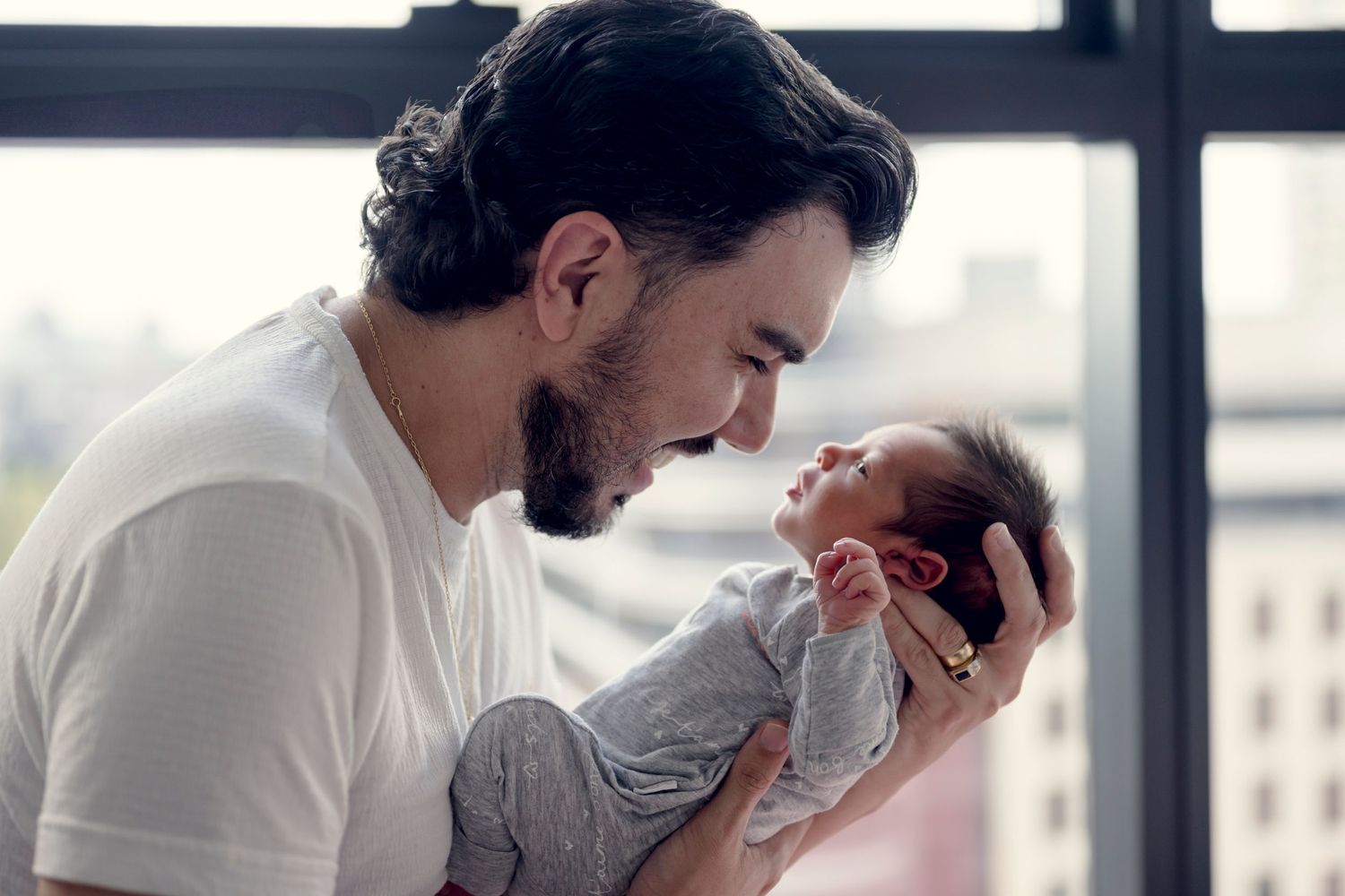 Father holding and gazing at newborn baby near a bright window.