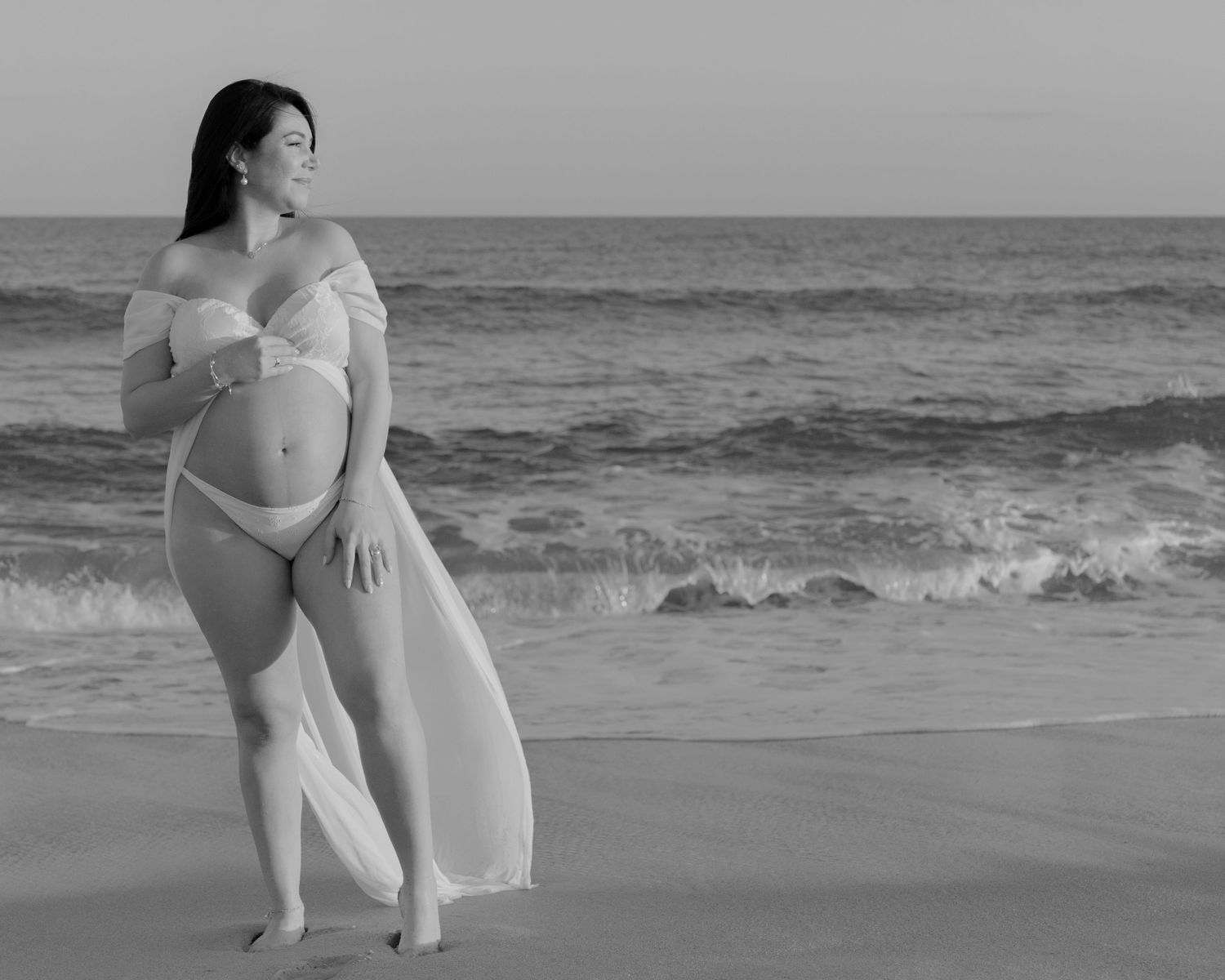 Artistic black and white maternity photo of an expectant mother in flowing fabric on a beach.
