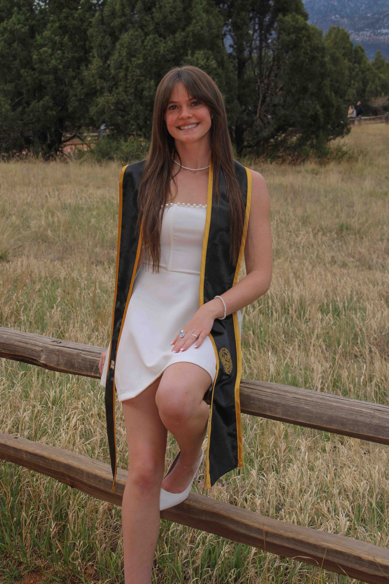 Smiling young woman in white dress and graduation stole posing by rustic fence in scenic meadow.
