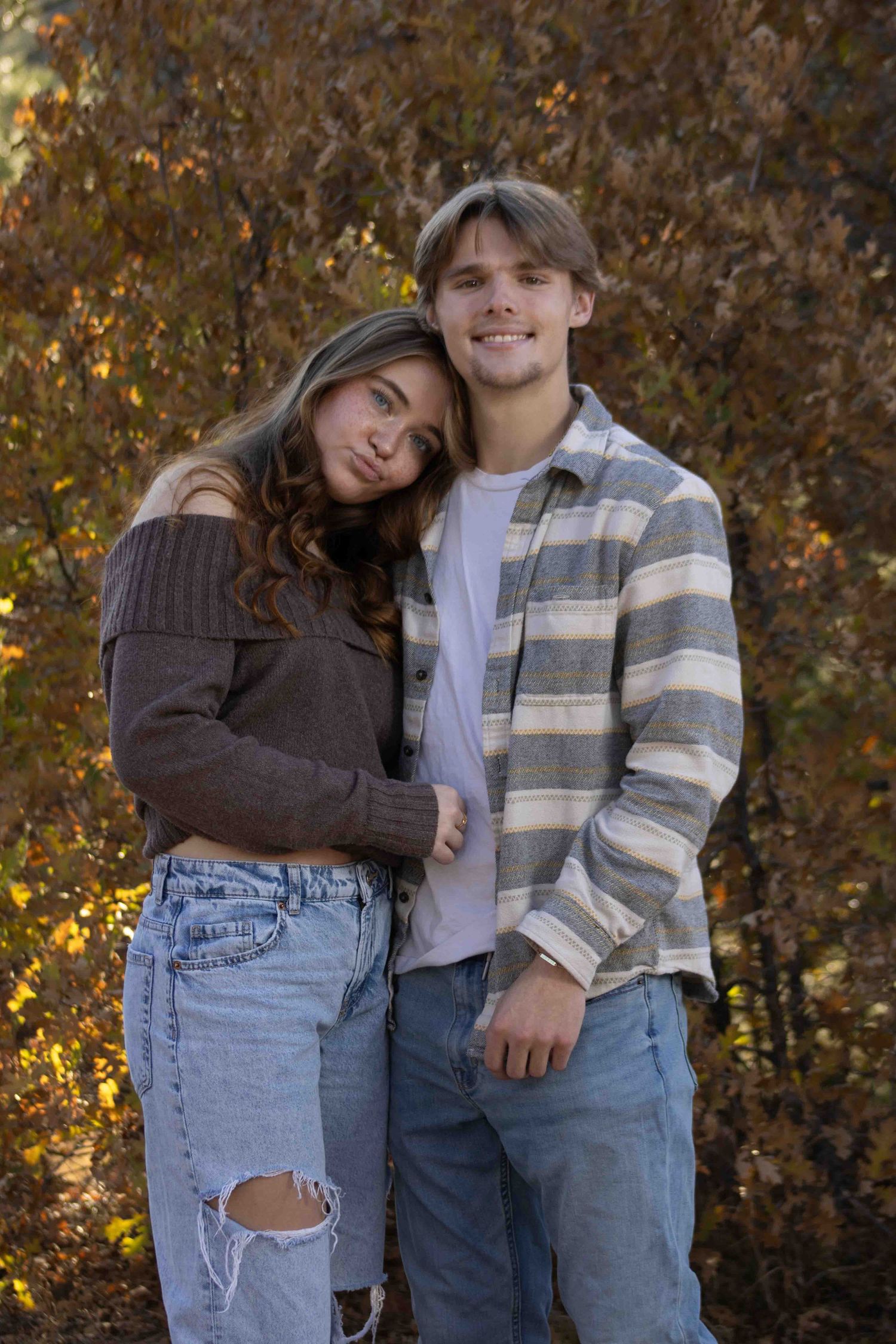A couple in matching brown and striped outfits poses together against fall foliage.