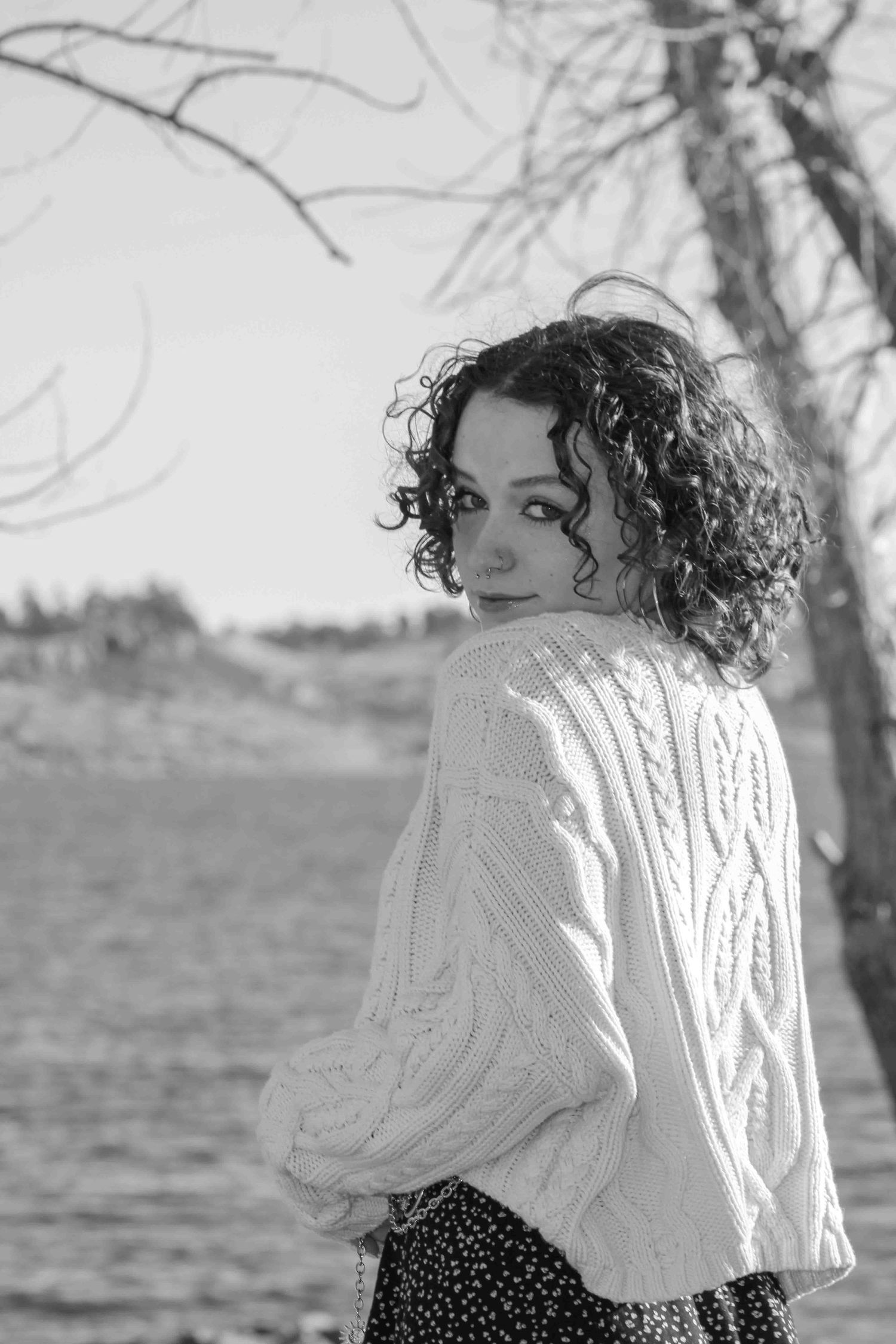 Person in white cable knit sweater poses by water on overcast day in vintage style portraits.