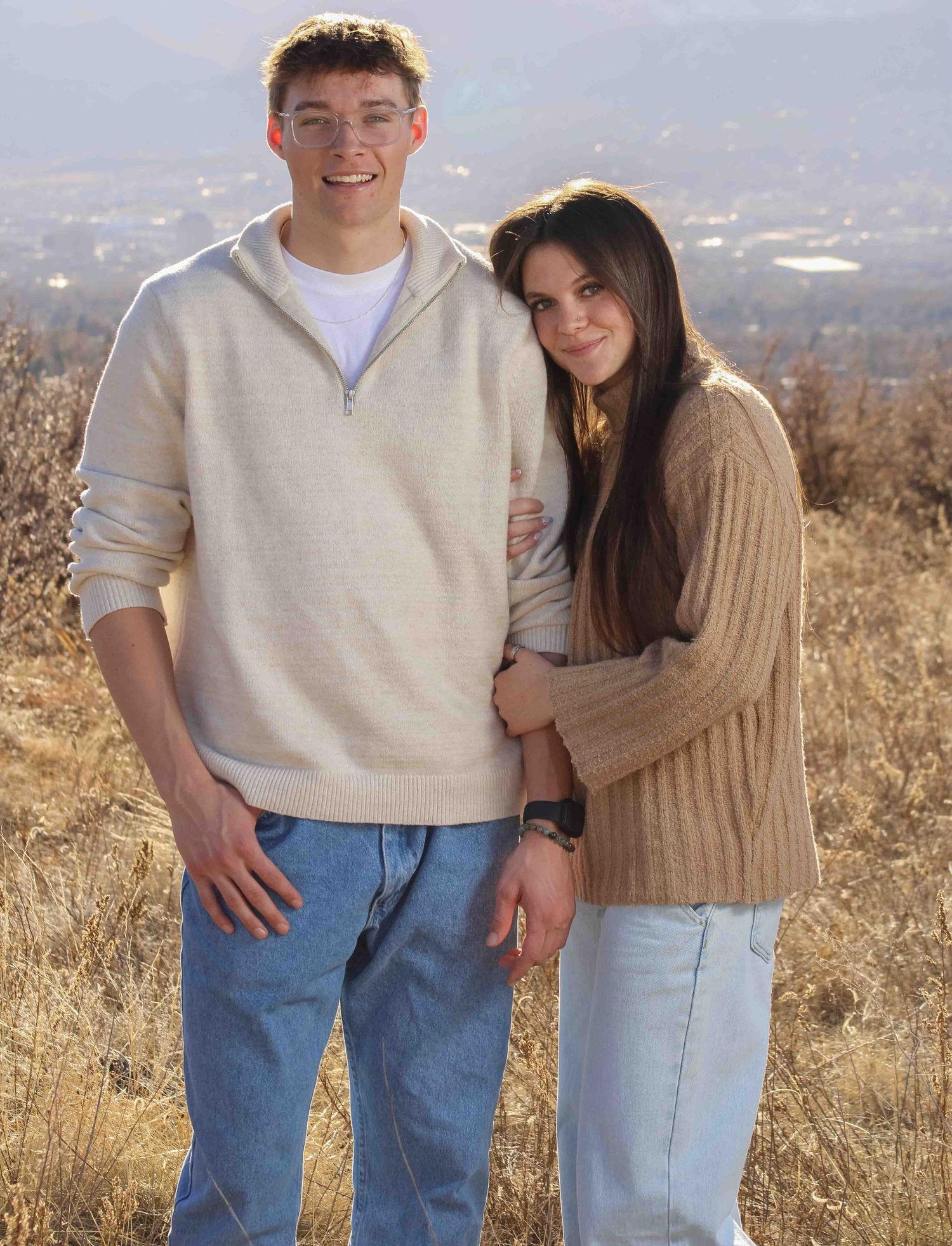 Couple in coordinating neutral sweaters poses together on hillside during golden hour.