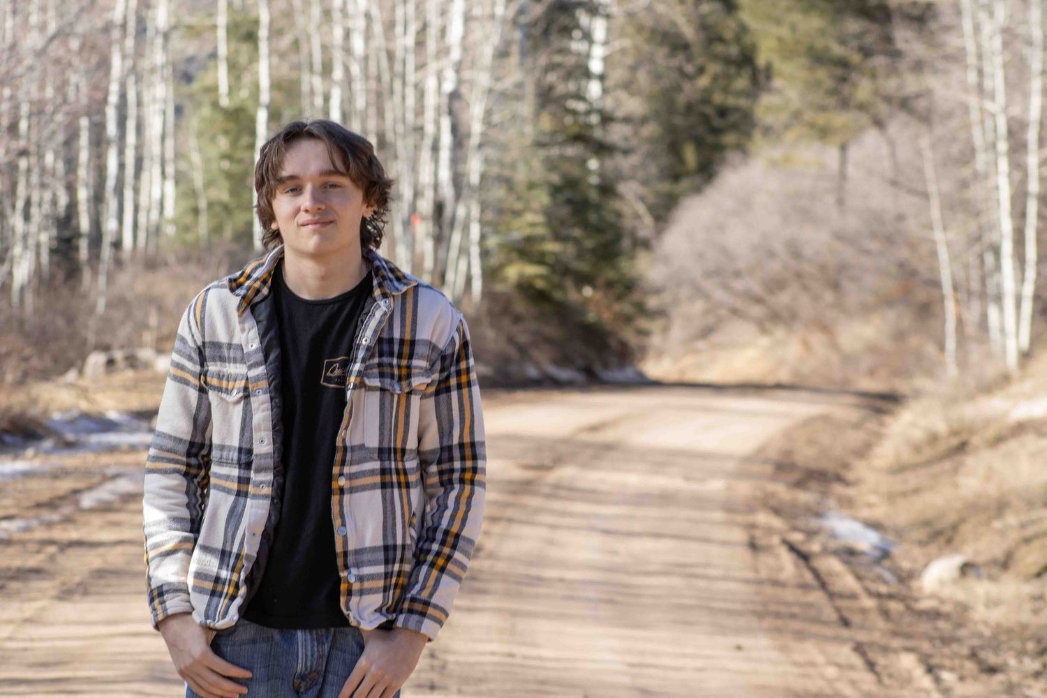 A person wearing a plaid flannel jacket stands on a dirt road with birch trees in the background.
