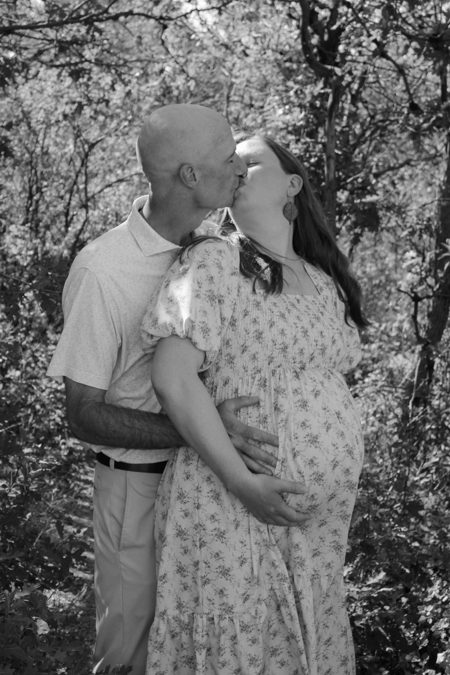 Black and white maternity photo of a couple kissing outdoors, man holding pregnant woman's belly among blooming trees.