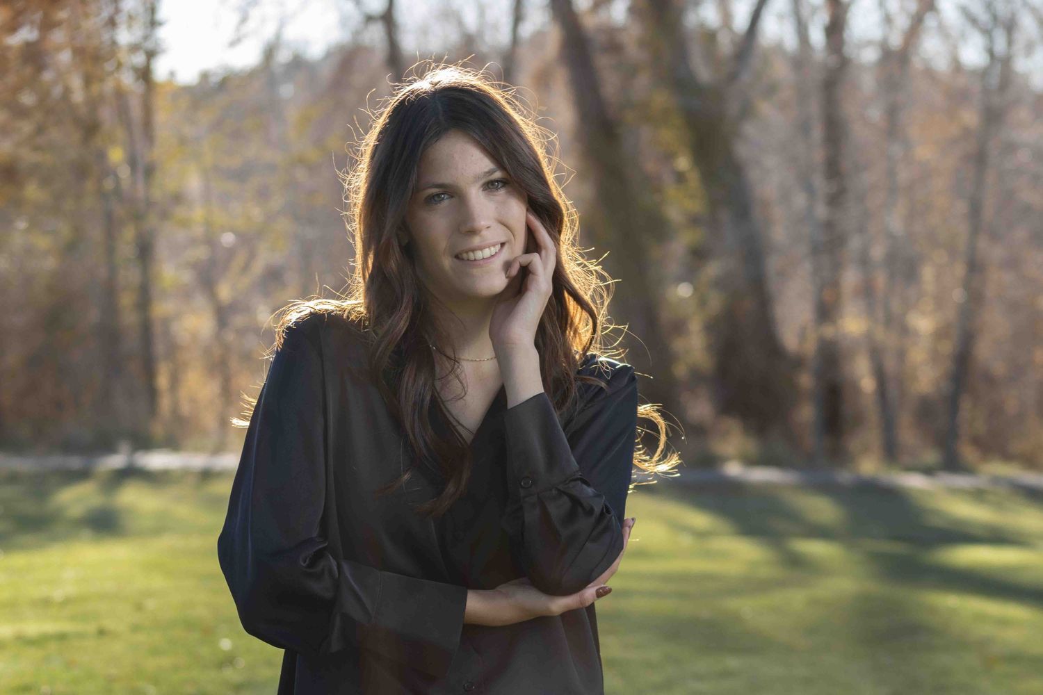 A person in a black sweater poses outdoors in an autumn setting with sunlit trees in the background.
