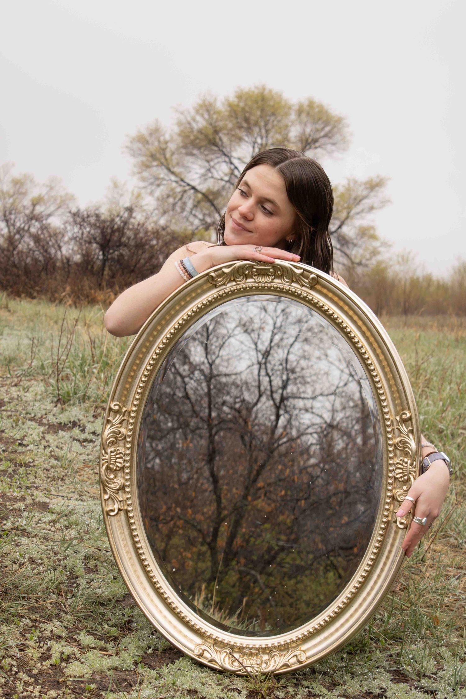 An ornate gold vintage mirror reflects bare trees in an autumn field on a cloudy day.