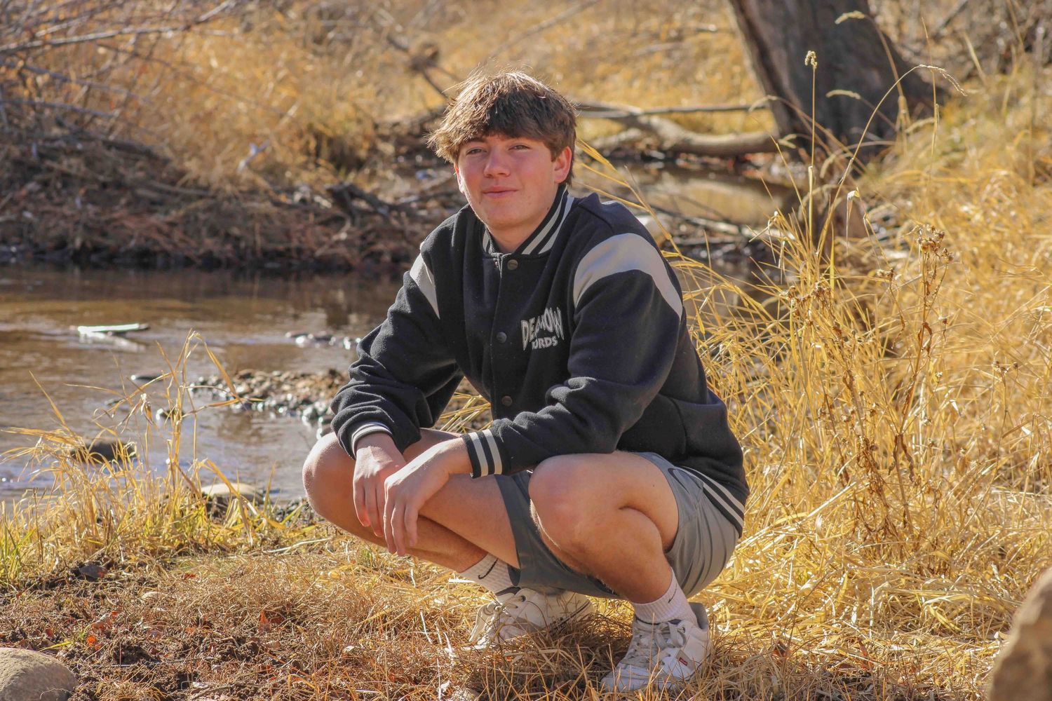A person in a hoodie crouches near a stream bank with dry grass and autumn scenery.