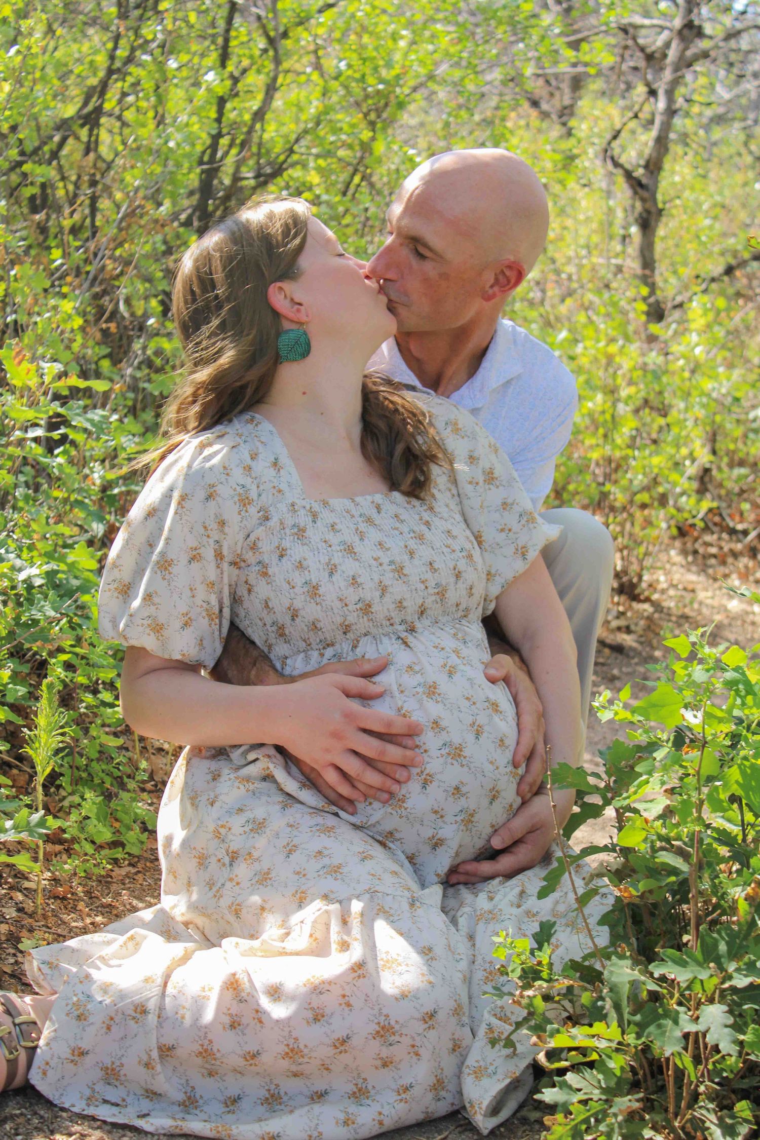 Pregnant woman in floral dress sits outdoors as bald man kisses her cheek, both cradling her baby bump.