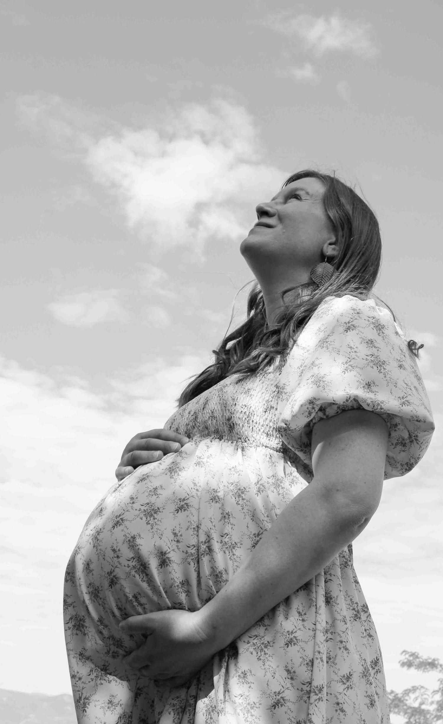 Black and white photo of a smiling pregnant woman in a floral dress cradling her belly while looking up at a cloudy sky.