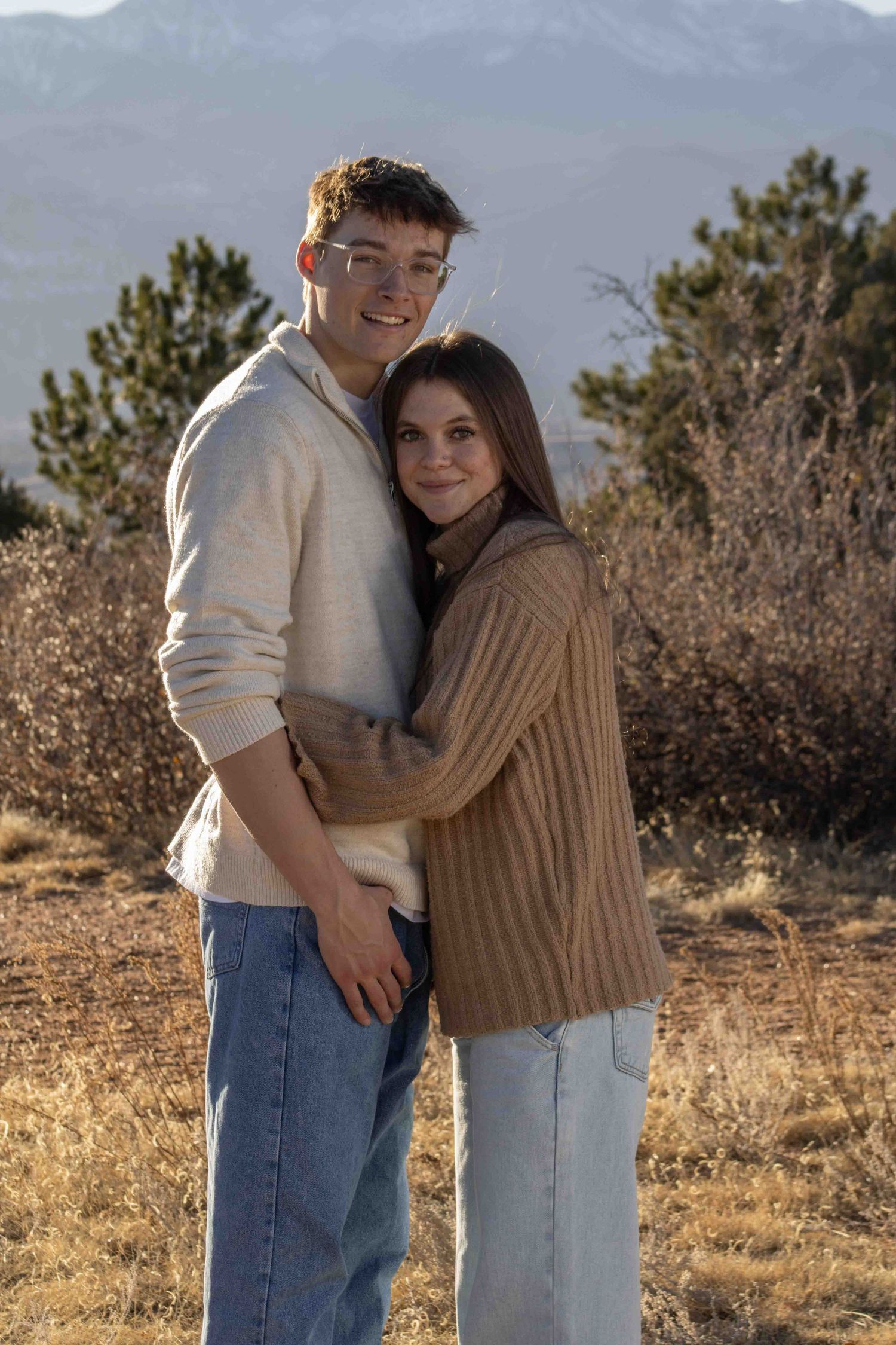 Couple in neutral sweaters poses affectionately against mountain landscape at sunset.