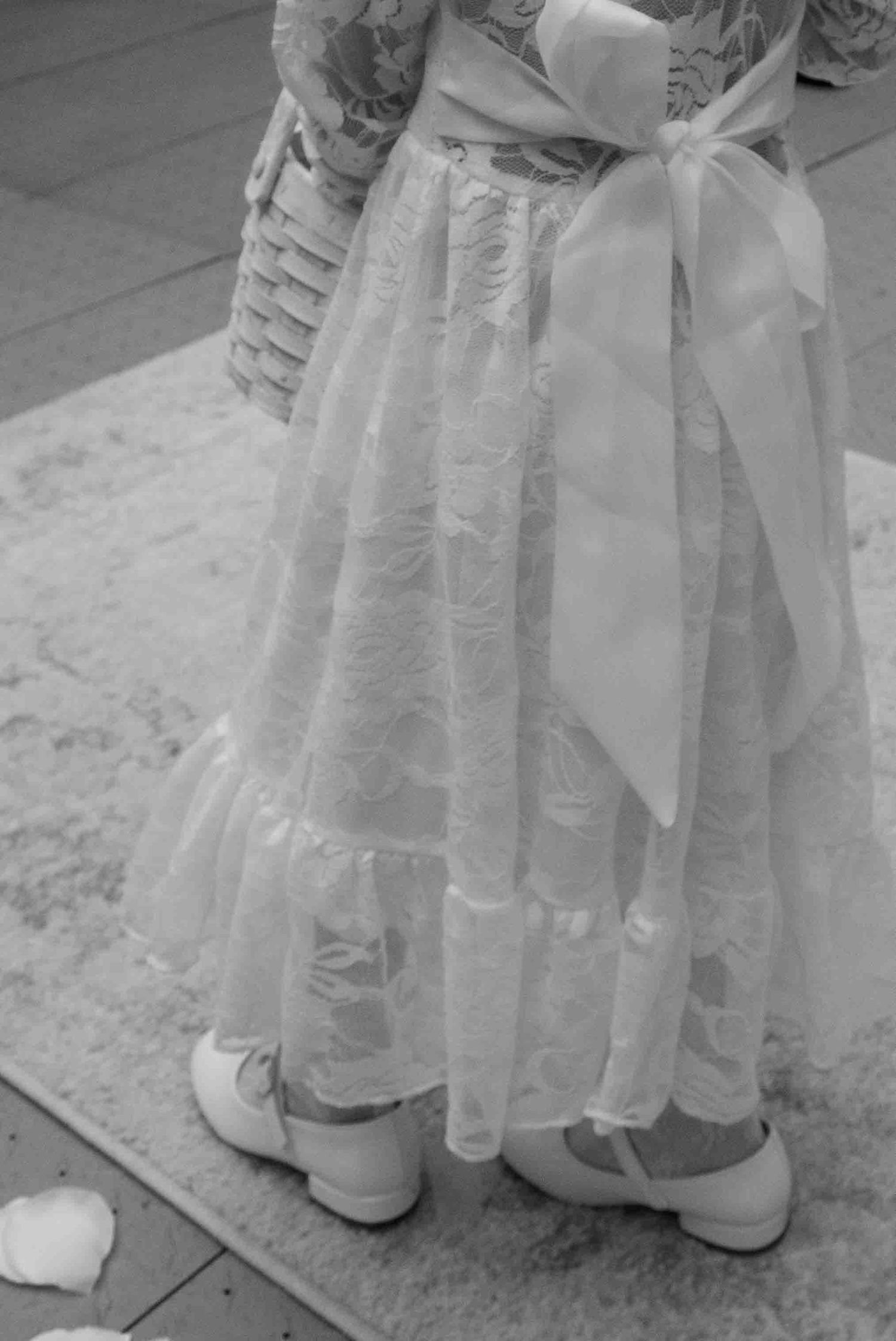 Black and white photo of a flower girl in a white lace dress with bow, holding a basket, wearing white shoes.