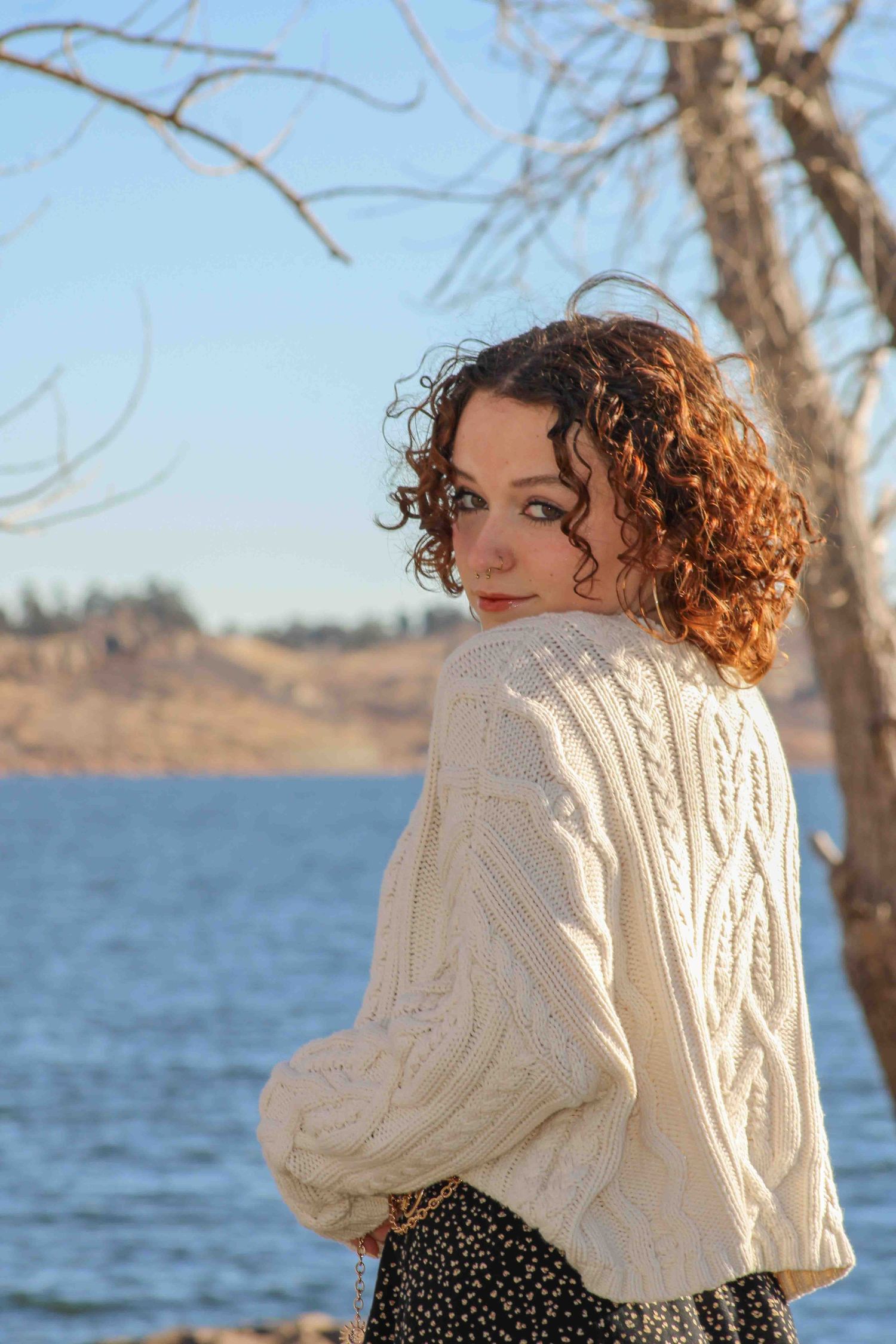 Figure wearing a cream cable knit sweater poses by a lake on a sunny day.